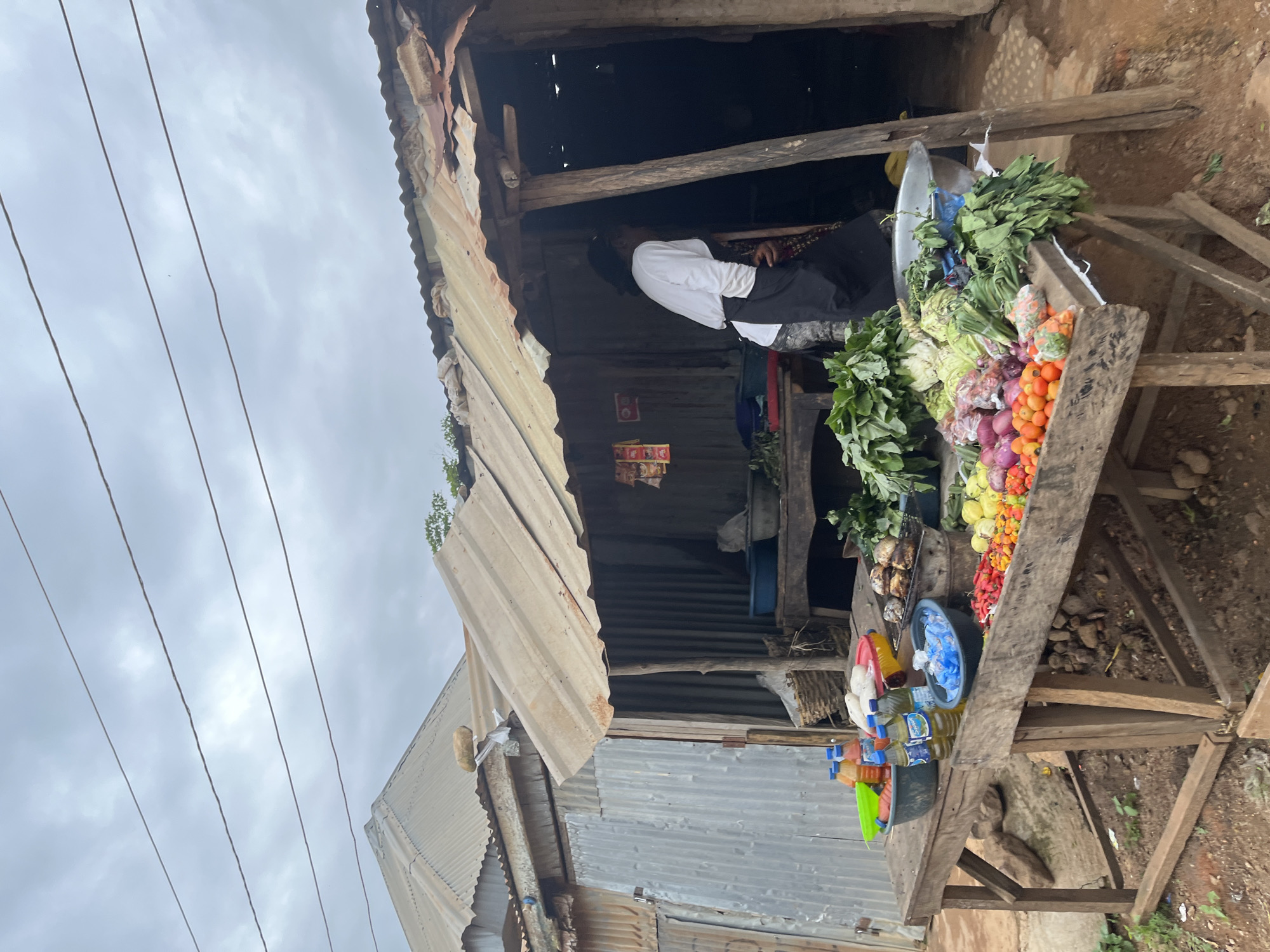 The image depicts an outdoor market stall in Jos, Nigeria, under a cloudy sky. The stall, constructed from corrugated metal and wood, displays a variety of fresh produce on a wooden table. Items for sale include leafy greens, onions, tomatoes, peppers, and what appear to be some root vegetables or fruits. There are also several bottles of beverages and oil on the table, along with some packaged goods and a basket. A woman, wearing a white long-sleeved shirt and a dark apron, is partially visible inside the stall, attending to the goods. The ground is dirt, and there is evidence of rough construction in the surroundings, with corrugated metal siding on adjacent structures and a rustic wooden support for the stall. The overall scene suggests a local, informal marketplace.