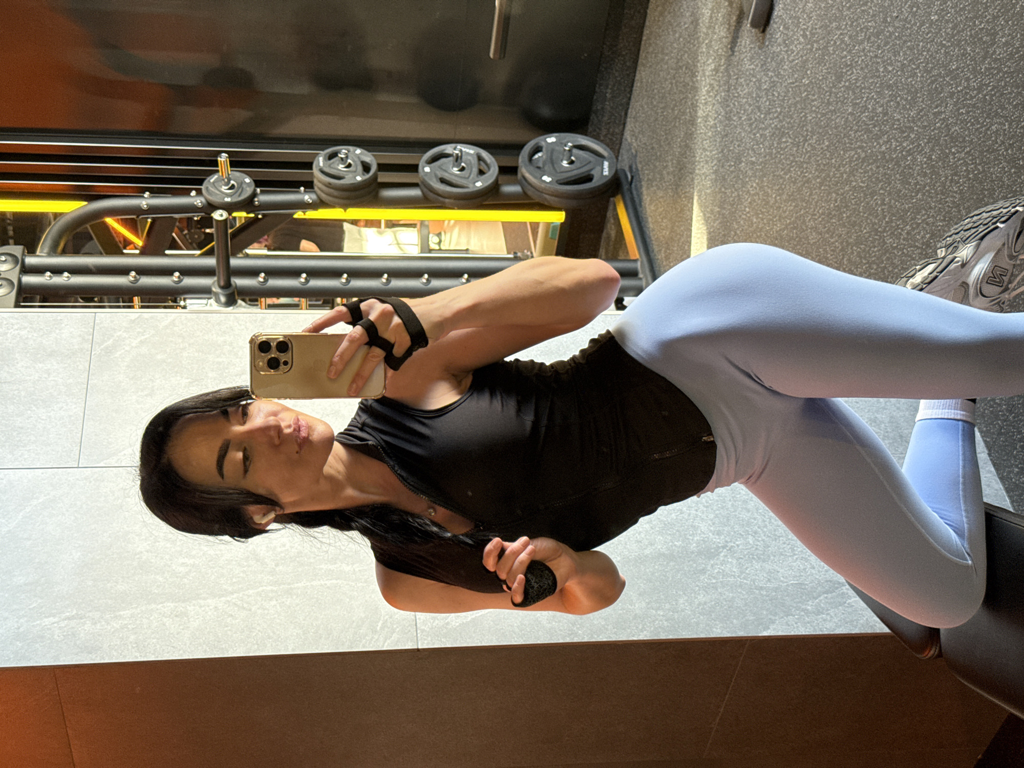 A woman is taking a selfie in what appears to be a gym. She is wearing a black tank top and light blue leggings with athletic shoes. The gym has weightlifting equipment in the background. There is no information to identify the city of Krasnoyarsk, Russia in the image.