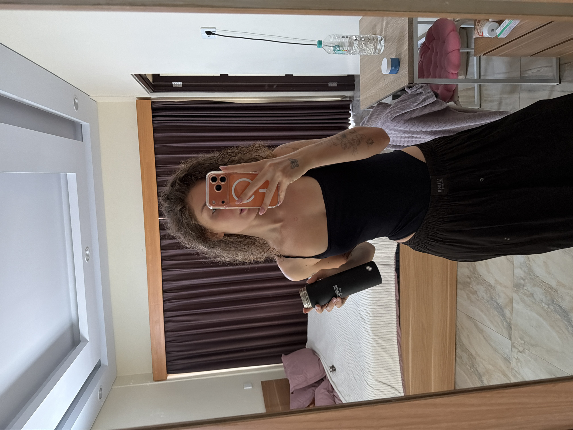 A person is taking a mirror selfie in what appears to be a bedroom or hotel room. The person is holding a black water bottle in their left hand and an orange-cased smartphone in their right hand, which is being used to capture the reflection. They are wearing a black tank top and dark, loose-fitting pants. The room features a bed with a white and light-colored striped comforter and several pink pillows, a wooden headboard, and dark brown curtains. To the right, a wooden table holds a plastic bottle of water, a small white jar with a blue lid, and a pink cushion on a chair. The floor is tiled with a marble-like pattern. The lighting suggests it is daytime. The location is specified as North Kuta, Indonesia.