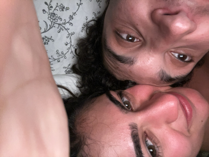 A close-up shot of two people lying in bed. The woman is on the bottom with curly hair and the man is on top with curly hair. The woman has brown eyes and defined eyebrows. The man also has brown eyes. They are close together, with the woman's face slightly turned toward the camera. A floral patterned pillow or blanket is visible in the background. The shot is taken at an angle, giving a slightly tilted perspective of their faces.