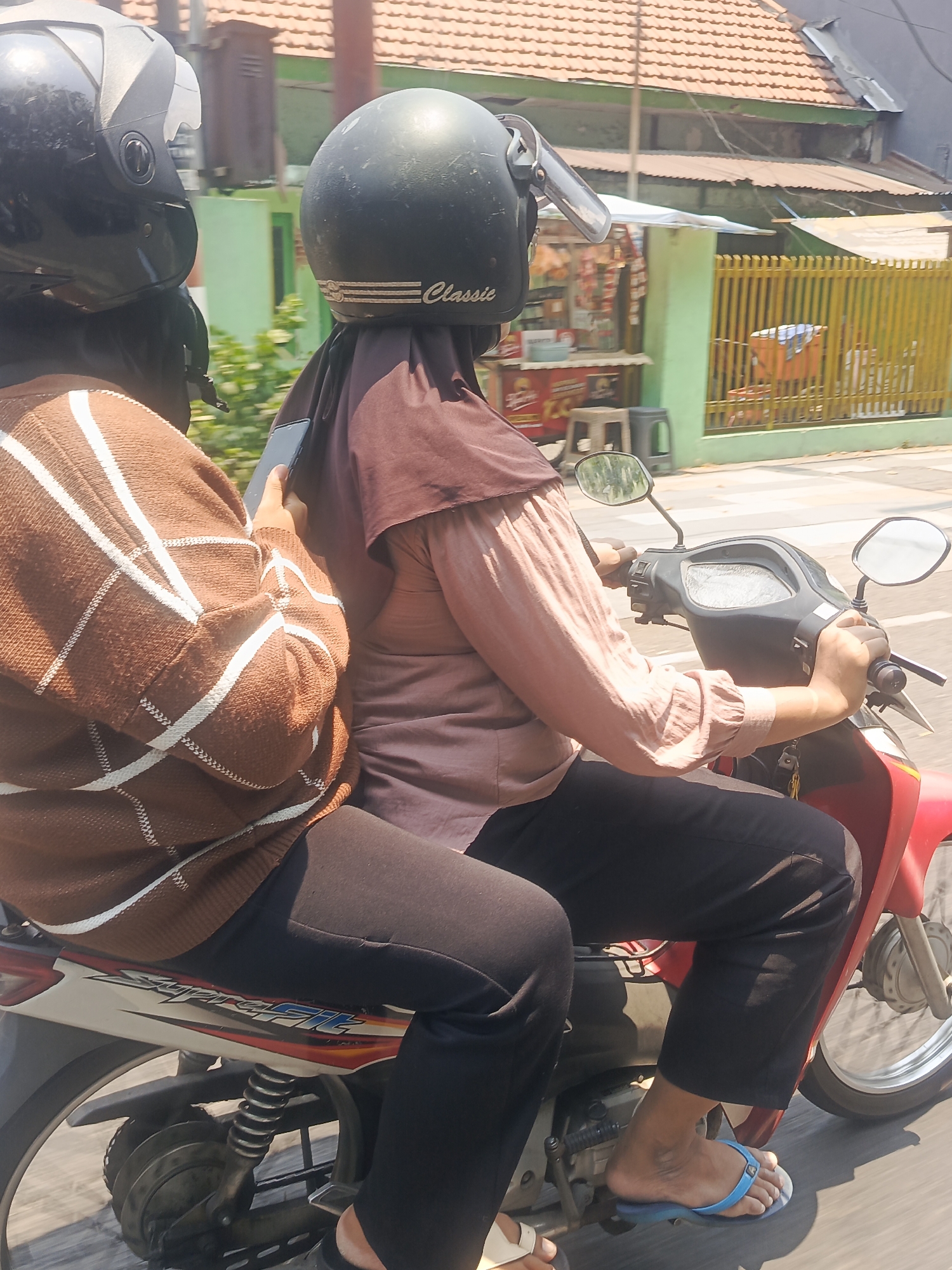 Two individuals are riding a red motorcycle in Surabaya, Indonesia. The person in the front, driving the motorcycle, is wearing a maroon hijab and a light pink long-sleeved shirt. They are holding the handlebars and appear to be looking forward. The person in the back is wearing a brown sweater with white stripes and black pants. They are holding a smartphone in their left hand. The motorcycle is moving on a paved road with a pedestrian crossing visible. In the background, there are buildings with tiled roofs, a green fence, and a street vendor stall. The scene is illuminated by bright daylight, suggesting it is daytime.