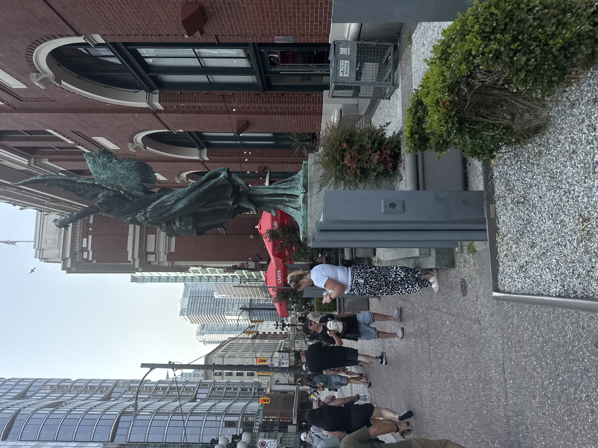 The image captures a street scene in Vancouver, Canada, likely in the Gastown or Downtown area, characterized by a mix of historic brick architecture and modern glass skyscrapers. In the foreground, a weathered bronze statue of an angel with outstretched wings stands tall. Adjacent to the statue is the brick facade of a building with large arched windows. A sidewalk paved with grey stones runs along the street, where several people are walking. Red umbrellas of an outdoor seating area are visible behind the statue. Further down the street, traffic lights and more contemporary buildings are apparent. The sky is overcast.