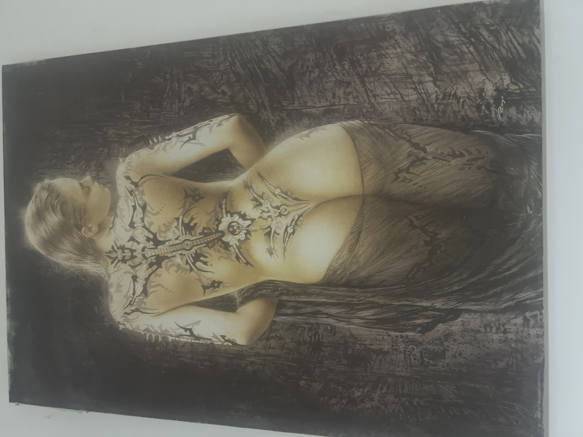 The image is a photograph of a painting that depicts a woman's back, from the neck down to her buttocks. She is facing away from the viewer, and her head is turned to the side, looking over her shoulder. The woman has blonde hair and is heavily tattooed with intricate, dark designs that cover her back, shoulders, and arms. She is draped in a dark, sheer fabric that covers her lower body and left arm. The background of the painting is dark and textured, with visible brushstrokes. The lighting in the painting is dramatic, with the woman's skin illuminated from the side, creating highlights and shadows. A signature, possibly "Royo", is visible in the lower right corner of the painting. The painting is hanging on a plain white wall, suggesting it is displayed in a home or gallery. There is no indication of the specific location within the city of Or Akiva, Israel.
