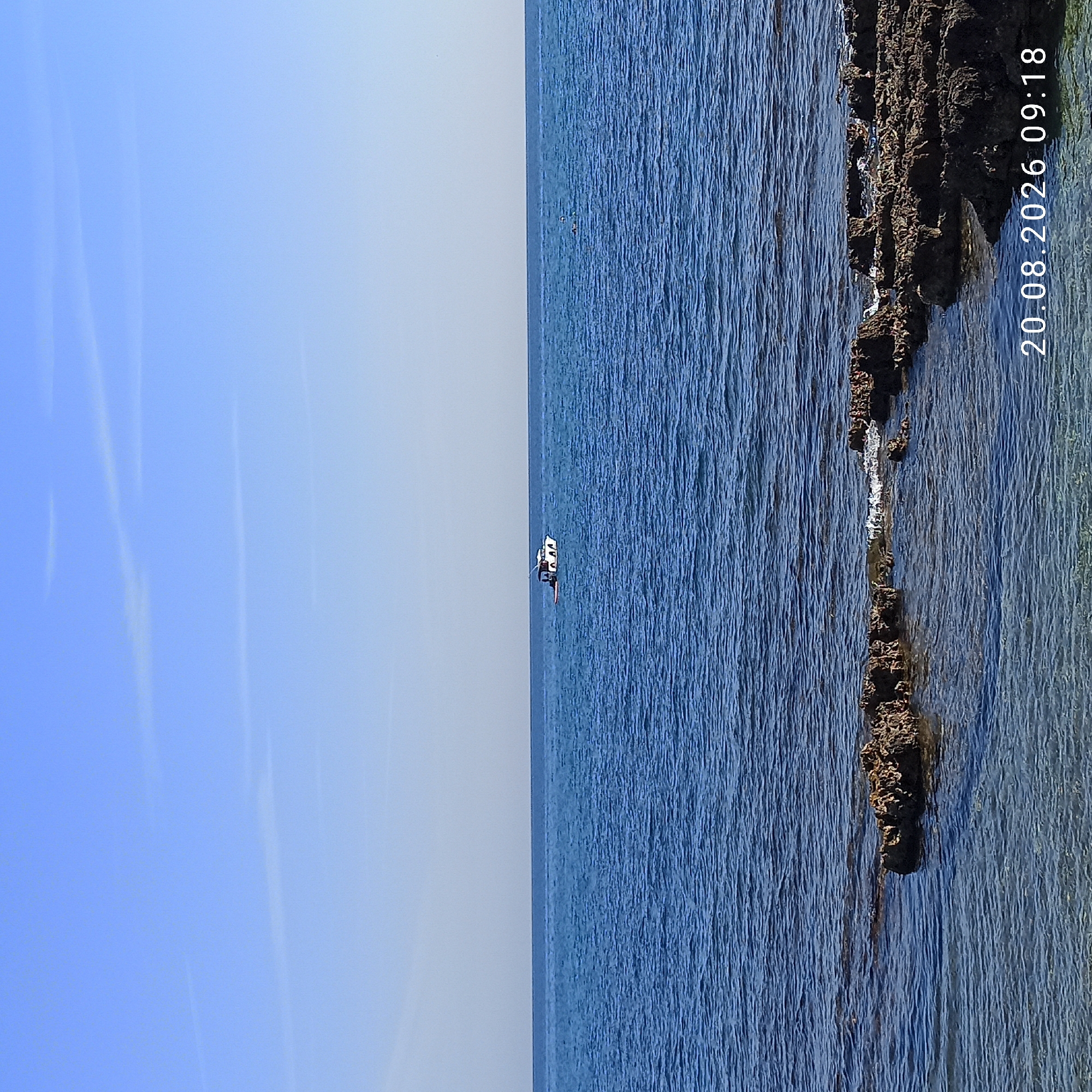 The image depicts a calm, blue sea under a clear sky with a few wispy clouds. In the distance, a white boat with black trim is visible on the water. The foreground features dark, rocky shorelines partially submerged in clear, shallow water. The date stamp "20.08.2026 09:18" is present in the lower right corner. Based on the date and time, this is a recent image, and its location is likely Molfetta, Italy, a coastal city known for its scenic waterfront.