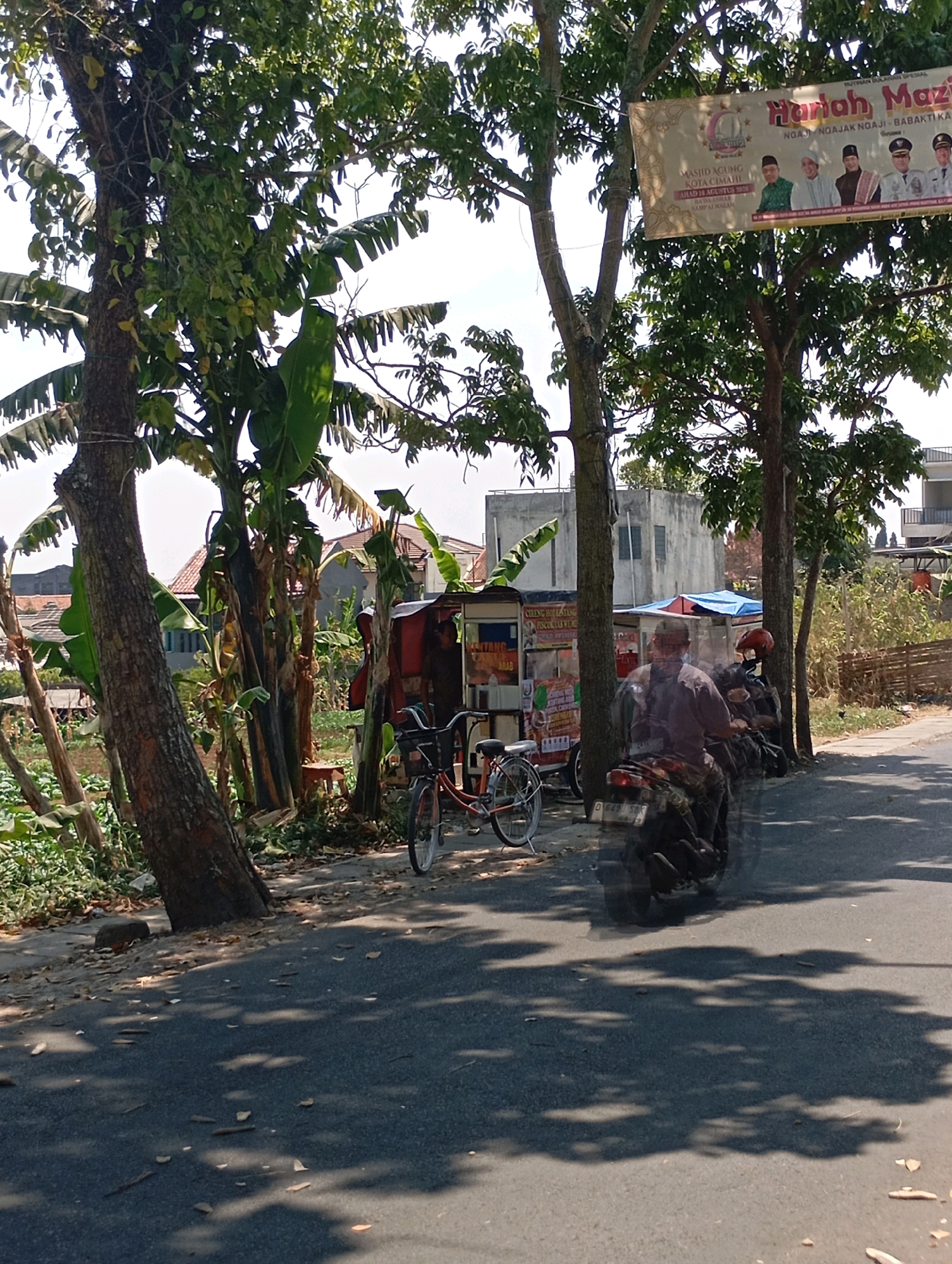 The image depicts a street scene in Cimahi, Indonesia, with a focus on a roadside stall and passing vehicles. A man wearing a dark shirt and a red helmet is riding a motorcycle in the foreground, partially obscuring a second motorcycle parked on the side of the road. To the left of the road, a small red and white food stall is visible, with a bicycle parked in front of it. Behind the stall, a few people are present, one appearing to be tending to the stall. The background features a building with a blue tarp on its roof and a banner advertising an event at Masjid Agung Kota Cimahi, an event that took place on August 15th, 2020. Lush green trees and foliage line the street, casting dappled shadows on the asphalt. The sky is bright and appears to be a clear day.
