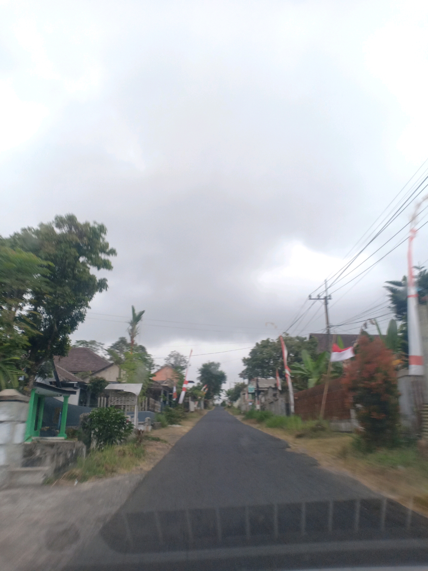 The image shows a paved road in the village of Kloposawit, Indonesia, with houses and trees lining the sides. The sky is overcast, with heavy clouds suggesting an impending storm or a generally humid climate. On the right side of the road, two Indonesian flags are prominently displayed, one fully unfurled and the other partially visible. These flags, with their red and white colors, indicate a national celebration or a display of patriotism. The houses appear to be traditional Indonesian dwellings, with some featuring red-tiled roofs. Vegetation is abundant, with lush green trees and plants adding to the tropical feel of the scene. The road is empty, with no people or vehicles visible, suggesting a quiet, perhaps early morning or late afternoon moment. The overall impression is that of a serene rural setting within an Indonesian village, with a touch of national pride represented by the flags.