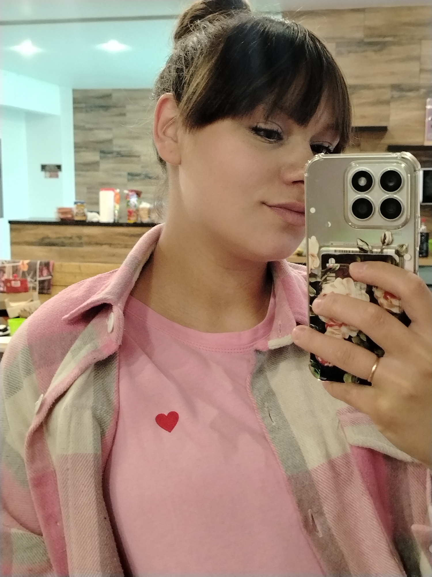 The image captures a partial view of a young woman taking a selfie indoors. Her face is shown in profile, with dark bangs framing her forehead and her hair styled in a bun. She has subtle eye makeup and a composed expression, gazing towards the smartphone she holds. She is wearing a light pink t-shirt featuring a small red heart on the left chest, layered beneath an unbuttoned pink, white, and grey plaid flannel shirt. A simple gold band is visible on her ring finger.
She holds a smartphone with a clear case that displays a floral pattern of white and pink roses on its back, partially obscured by her fingers. The phone's quad-camera system is prominently visible.
The background, which is blurred, suggests an indoor setting with bright artificial lighting. Details include light-colored walls, overhead light reflections on the ceiling, and a wooden counter or paneling with various containers and items, possibly indicating a cafe or shop environment. The activity depicts a casual self-portrait moment. This scene is set in what is understood to be Chaikovsky, Russia. There is no discernible text visible in the image.