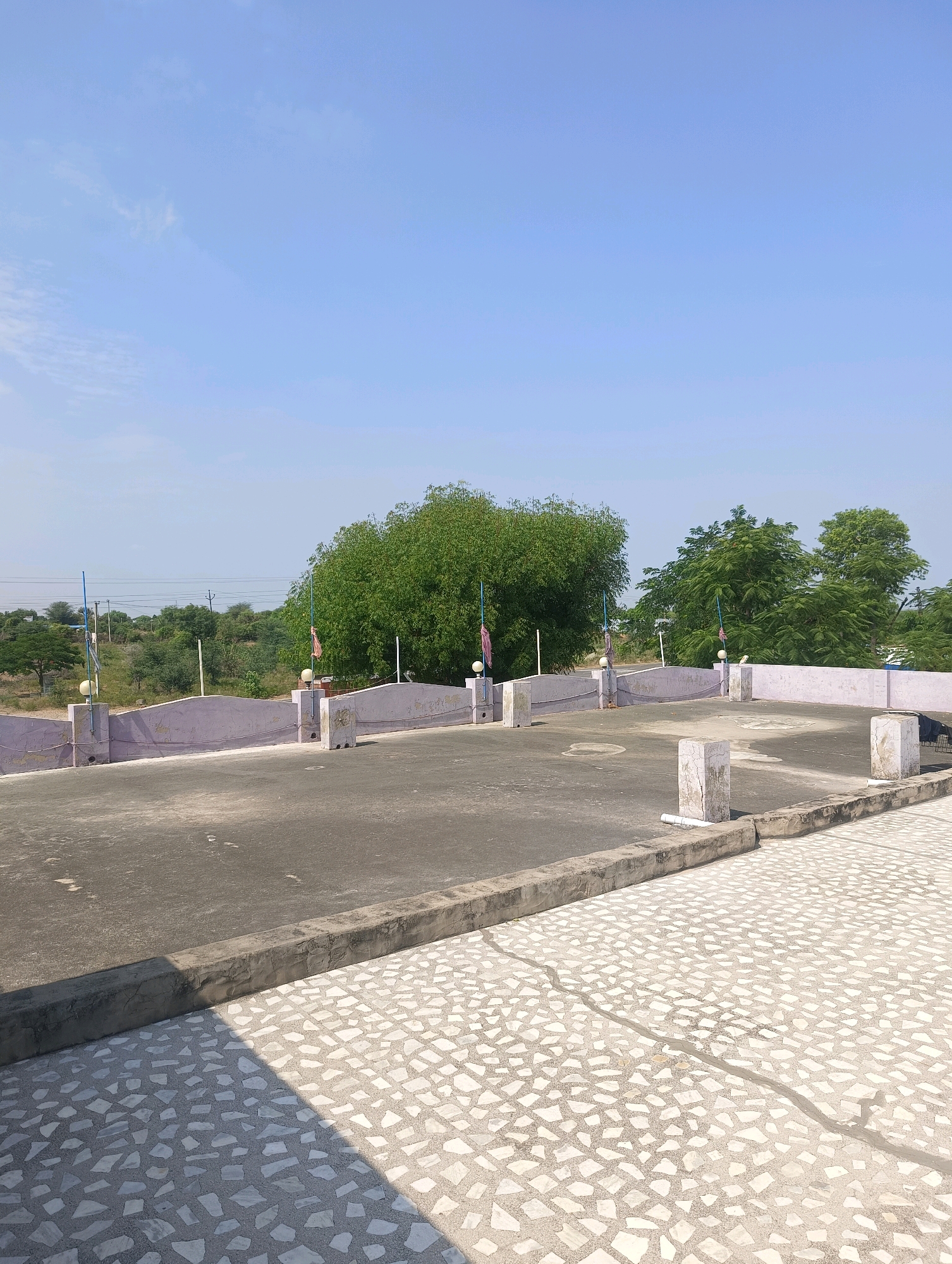The image shows a rooftop scene. The foreground features a textured floor made of white and gray irregular stone tiles, with a concrete border. To the left, a section of dark gray concrete pavement is visible, casting a shadow. The middle ground depicts a flat rooftop area with a light pink parapet wall. Several concrete pillars are present on the rooftop, some with flags attached. Beyond the rooftop, a landscape with green trees and vegetation is visible under a clear blue sky. Power lines are also seen in the distance. The scene appears to be a typical rooftop in a rural or semi-urban area. There are no people or animals visible in the image. The location is stated as Chhapri Bari, India.