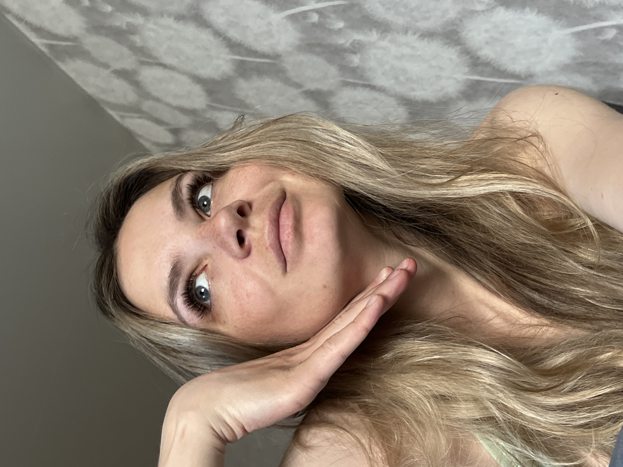 A young woman with blonde hair and blue eyes is posing indoors. She is resting her face on her hand, and her gaze is directed slightly upward. The background consists of a wall with a patterned wallpaper on the top and a plain, light gray wall on the bottom. The location is unknown, and there is no information indicating it's Chaikovsky, Russia.