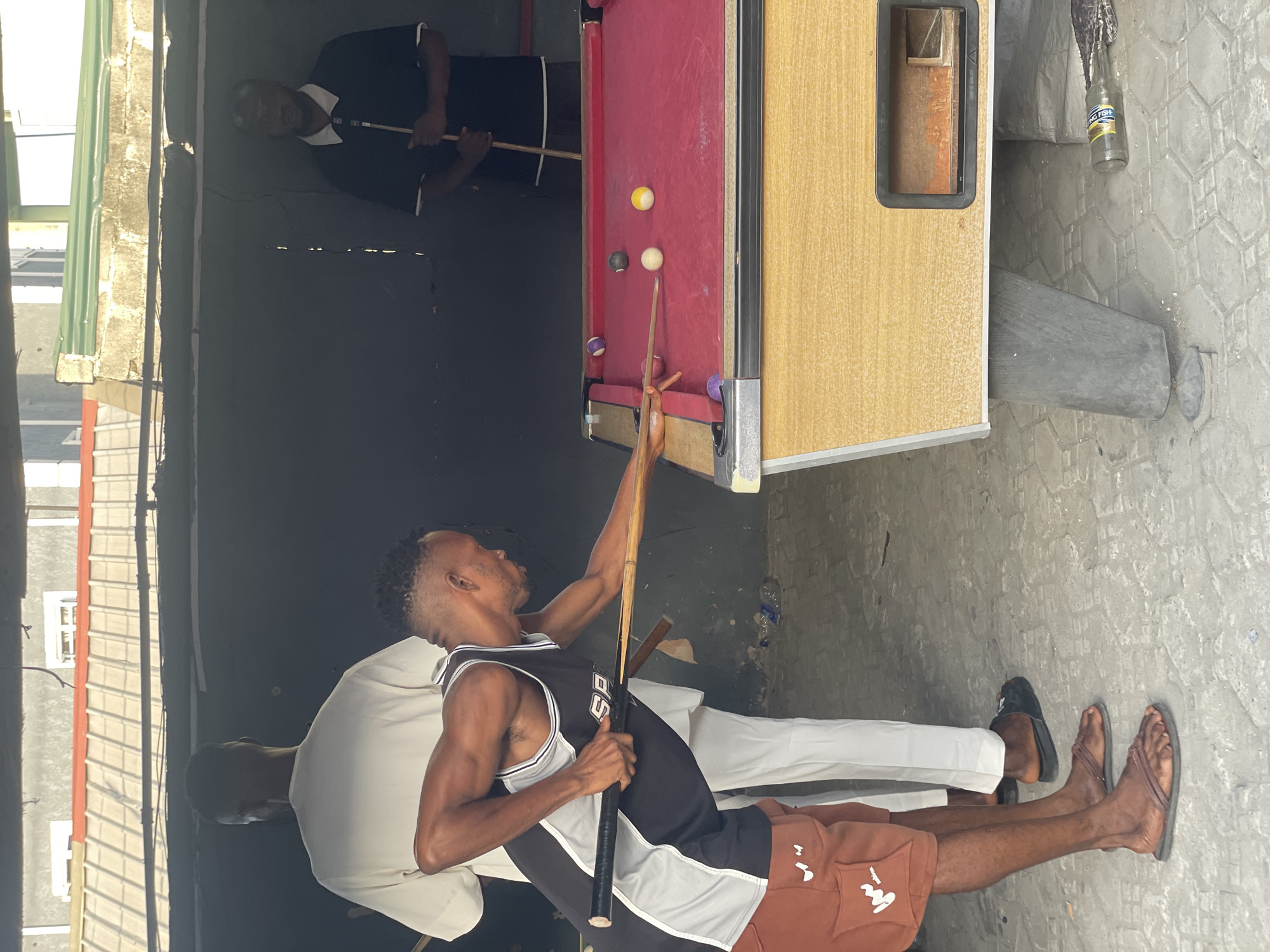 The image shows two men playing pool in what appears to be an outdoor establishment in Ado, Nigeria. One man in a Spurs jersey prepares to take a shot on a red pool table with several balls on it. Another man is seen standing at the other end of the table, holding a cue stick. The surroundings feature concrete flooring, a dark backdrop possibly indicating an enclosed space, and building structures visible in the periphery, suggesting an urban setting. Empty beer bottles are visible on the ground.