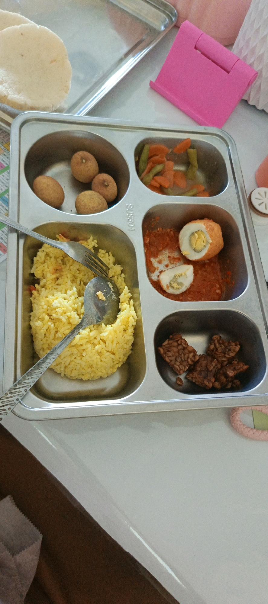 A metal tray with five compartments is positioned on a light-colored surface. The tray contains a meal. In the top left compartment are four small, oval, brown objects, possibly fried foods or fruits. To the right of these, in the top right compartment, is a portion of cooked vegetables, including sliced carrots and green beans, in a light liquid. The middle left compartment holds a generous serving of yellow rice, with a spoon and fork resting on it. To the right of the rice, in the middle right compartment, is a red sauce with two pieces of hard-boiled egg, one whole and one halved, which appear to be coated in an orange batter and served in the sauce. In the bottom compartment are several pieces of dark brown, rectangular-shaped food, likely tempeh. A pink tablet stand is visible in the background. The overall scene appears to be a meal being served in Desa Sarang Mandi, Indonesia.