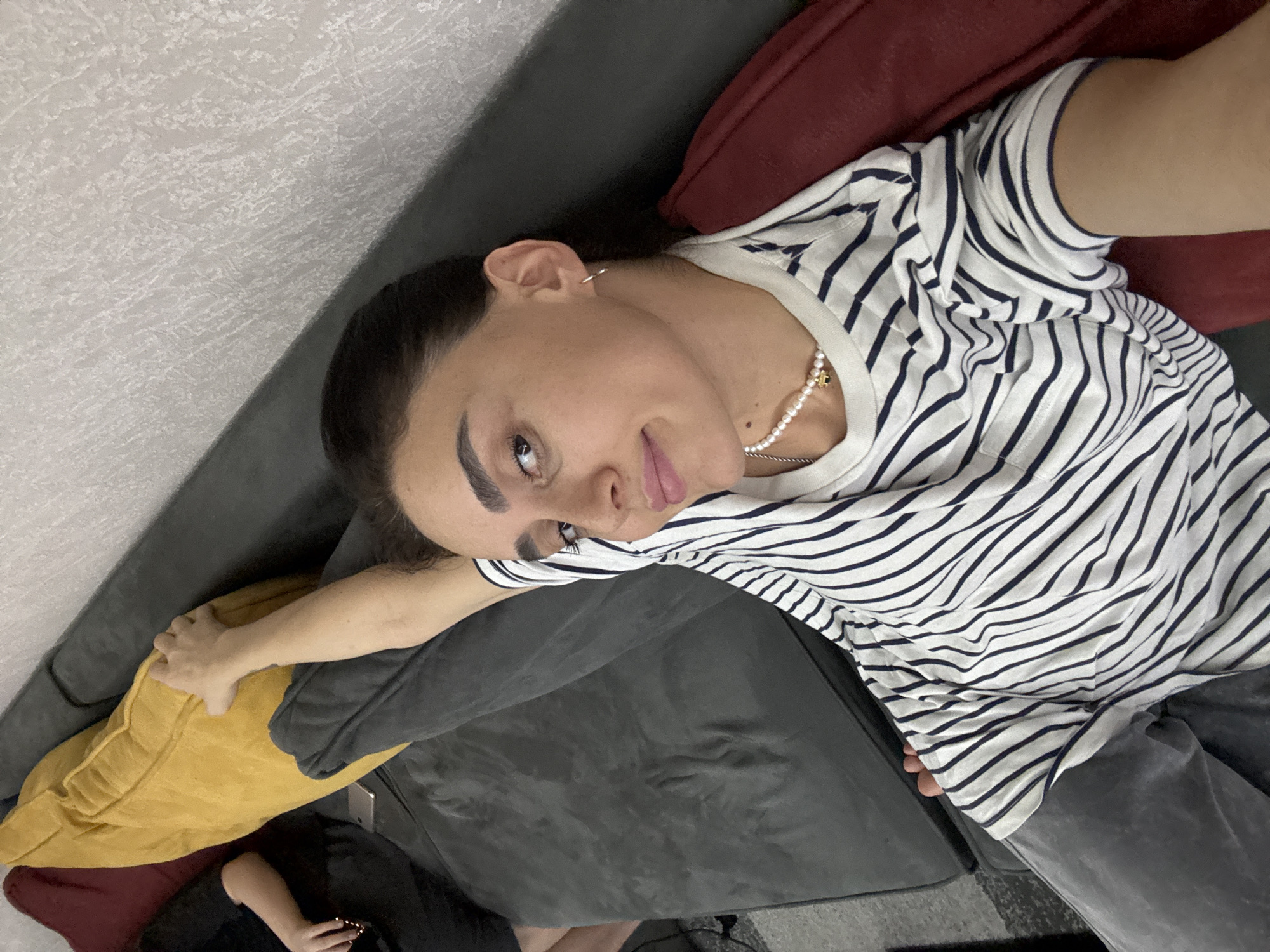 In an indoor setting, a woman with dark hair, styled back, is lying on a grey couch with a yellow cushion behind her head and a dark red cushion to her right. She is wearing a white t-shirt with navy blue stripes and grey pants. A pearl necklace with a gold charm is visible around her neck, and she has small hoop earrings in her ears. Her right arm is extended, resting on the couch. To her left, partially visible, is another person in a black shirt. The background wall has a textured, light-colored finish. No specific location in Tbilisi, Georgia, can be identified from the image.
