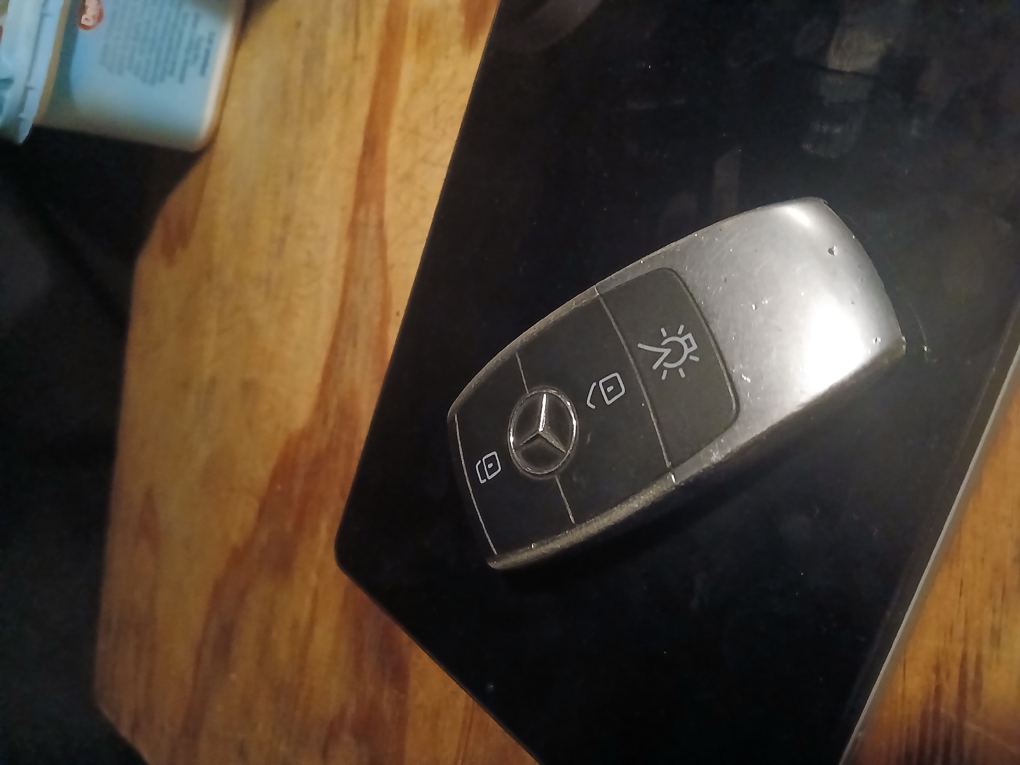 The image shows a Mercedes-Benz car key fob resting on a black surface, likely a tablet or laptop. The key fob is silver and black, with symbols for locking, unlocking, and a light. The surface it rests on is dark and reflective. In the background, a wooden surface with distinct grain patterns is visible, and a portion of a light-blue container, possibly food-related, is present. The overall lighting suggests an indoor setting, perhaps a home or office. The scene does not depict any specific location within the city of Douai, France, and contains no discernible people or actions.