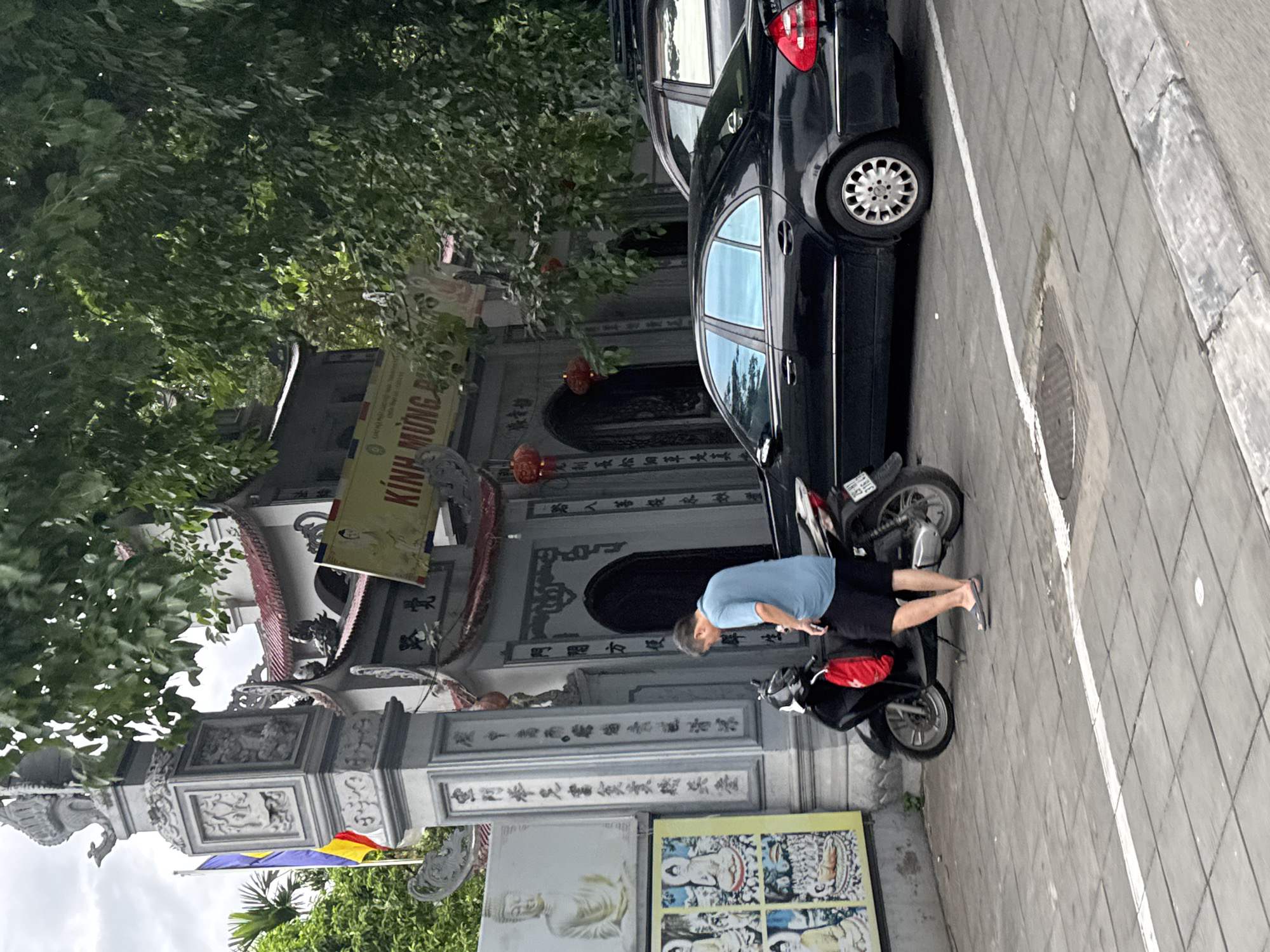 The image depicts a street scene in Hanoi, Vietnam, likely in front of a temple or pagoda. In the foreground, a man wearing a blue t-shirt and dark shorts stands next to a parked black motorcycle. He is facing downwards, appearing to interact with something on the motorcycle or on the ground. The motorcycle is black with silver accents and has a red bag hanging from the back. Behind the motorcycle, a black sedan is parked, with its rear visible. The car has tinted windows.
To the left of the man and motorcycle, a large, ornate stone pillar stands, intricately carved with figures and text in what appears to be Vietnamese. To the left of the pillar is a colorful display of Buddhist artwork or imagery, featuring multiple depictions of Buddha. Beyond the pillar and the man, a building with a traditional tiled roof and arched doorways is visible, suggesting it is a place of worship. A banner with Vietnamese text hangs above the entrance. Red lanterns are suspended from the building.
The ground is paved with square tiles, and a white line is painted on the pavement. Lush green trees with dense foliage overhang the scene, providing shade and contributing to the overall atmosphere. The sky is overcast. The architectural style and the presence of Buddhist imagery strongly suggest a location within Hanoi, Vietnam.