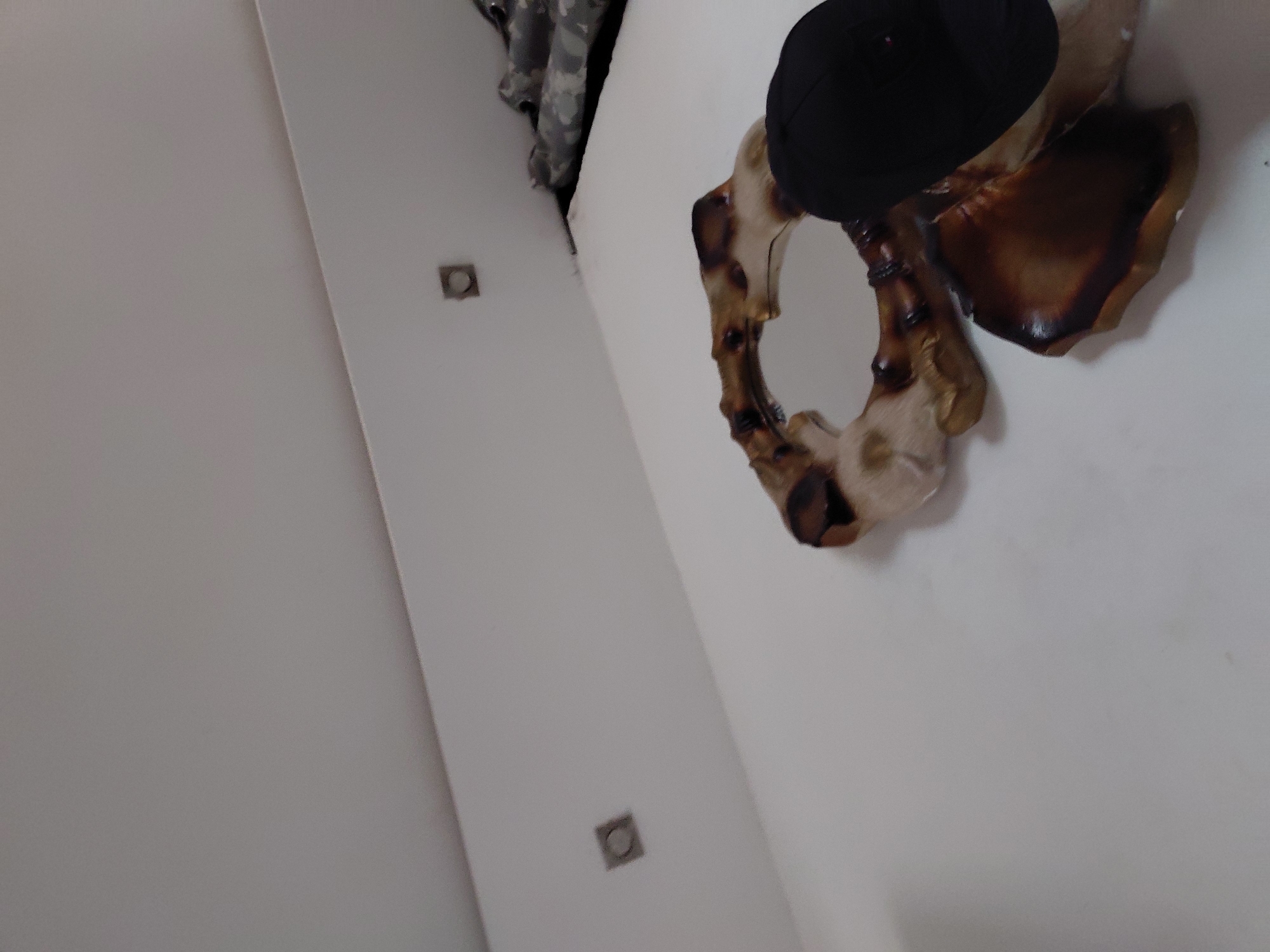 The image, captured in Santa Ana, Venezuela, displays an indoor scene from a rotated perspective, likely looking upwards at a white wall. The primary subject is an organically shaped mirror or decorative frame mounted on the wall. This piece appears to be made from wood or a similar natural material, exhibiting a variety of rich brown, reddish-brown, and lighter, creamy tones, with a polished finish that accentuates its texture. An irregularly shaped reflective surface is visible within the frame, confirming it as a mirror. A solid black cap or hat rests on the upper part of this decorative object.
Further details on the white wall include two small, square fixtures with dark or metallic rims, positioned vertically along the wall. These could be electrical outlets or recessed lighting elements. In the upper left corner of the frame, a fragment of camouflage-patterned fabric is partially visible. The scene is static, depicting a decorated interior space. No specific activity is occurring, and there is no visible text. The lighting suggests an indoor setting, but the time of day is not discernible.