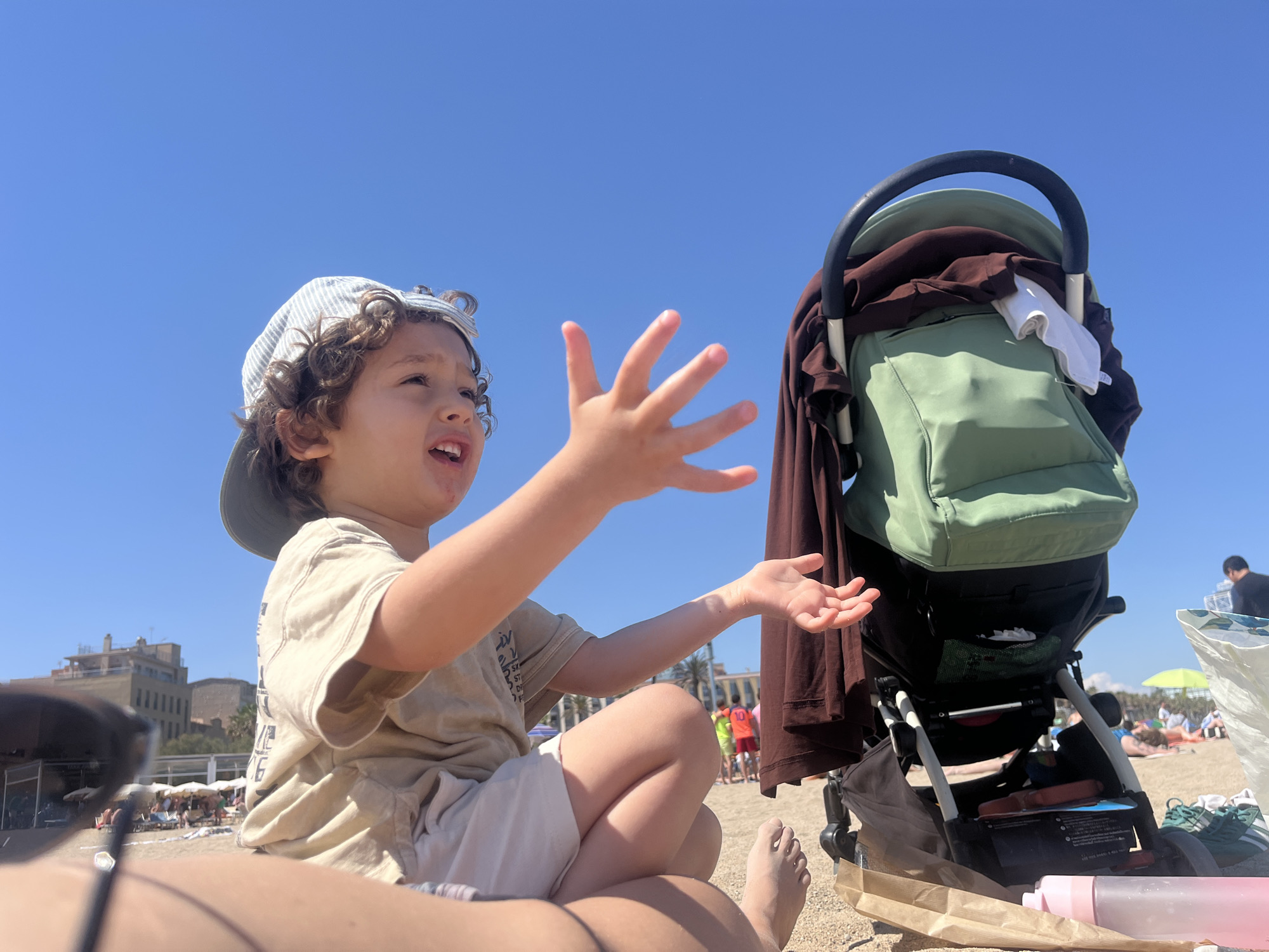 The photograph captures a sunny day at a beach in Barcelona, Spain. A young child with curly hair and a striped hat sits on someone's lap, their hands outstretched as if playing or gesturing. The child wears a light-colored t-shirt and shorts. Beside them, a green baby stroller holds a brown garment and a white cloth. The beach is populated with people sunbathing and relaxing, and buildings are visible in the background. The sky is a clear blue, indicating good weather. Sand is visible on the ground.