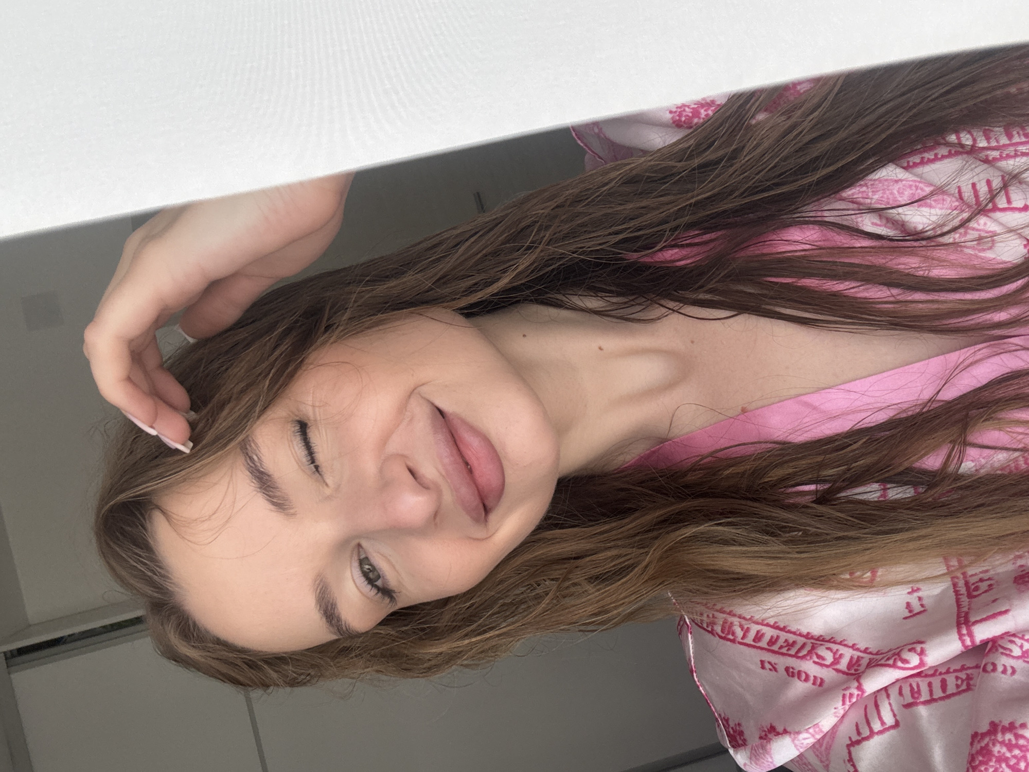 A young woman with long brown hair is positioned close to the camera. She is wearing a pink and white patterned robe and winking with her left eye. Her hair is partially wet. She is holding her right hand near her head, and a corner of a white object is in the upper left of the frame. The background is blurred, but it appears to be an indoor setting. There is no information provided in the image to determine its location in the city of Budva, Montenegro.