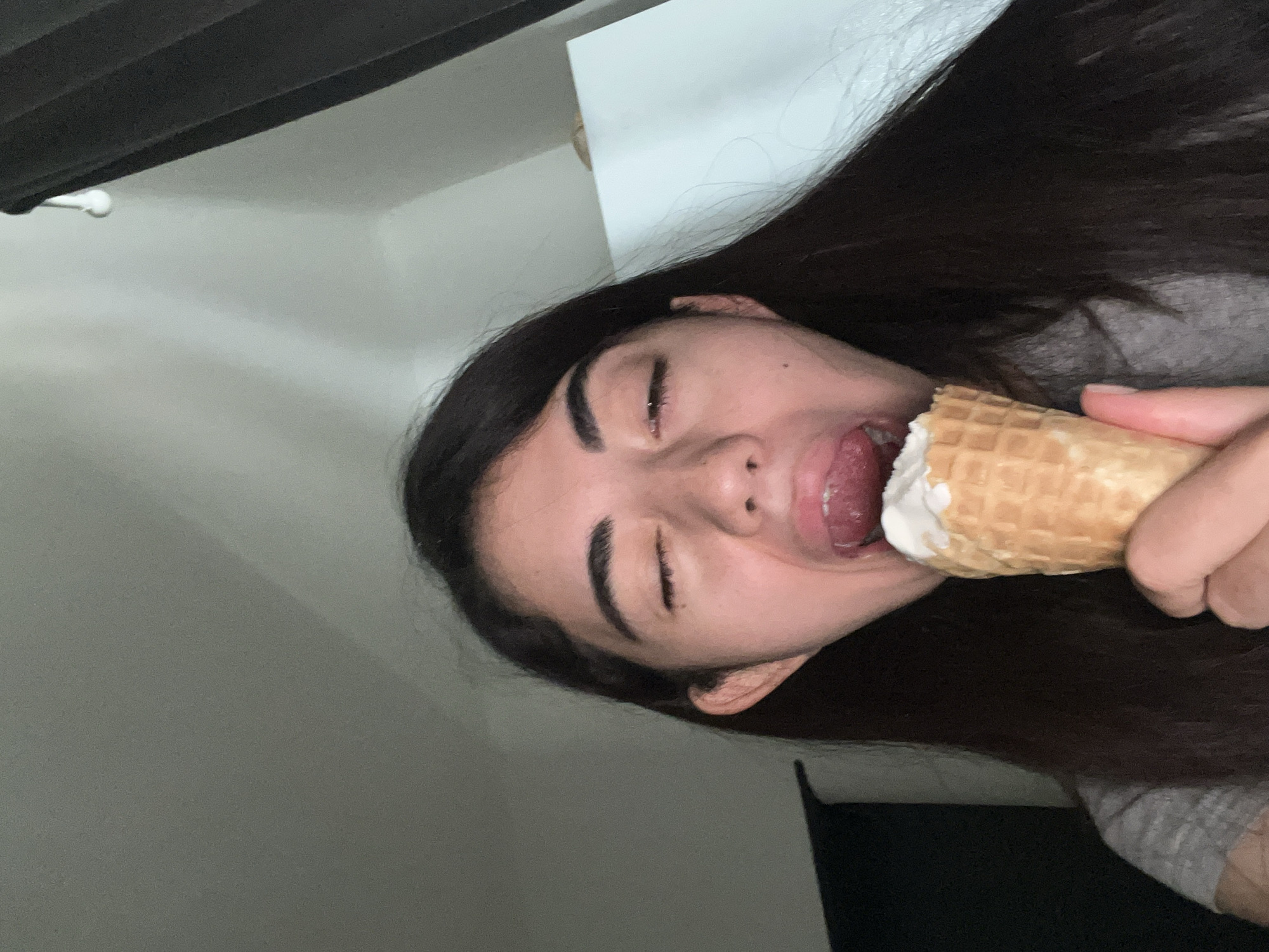A young woman with long, dark hair is shown from the chest up, with her eyes closed and her mouth open, about to eat an ice cream cone. She is wearing a gray t-shirt. The background consists of a gray wall and a white surface, possibly a cabinet, with dark curtains to the right. The lighting is dim, suggesting an indoor setting. The location in Chotěšov, Czechia is not discernible from the image.