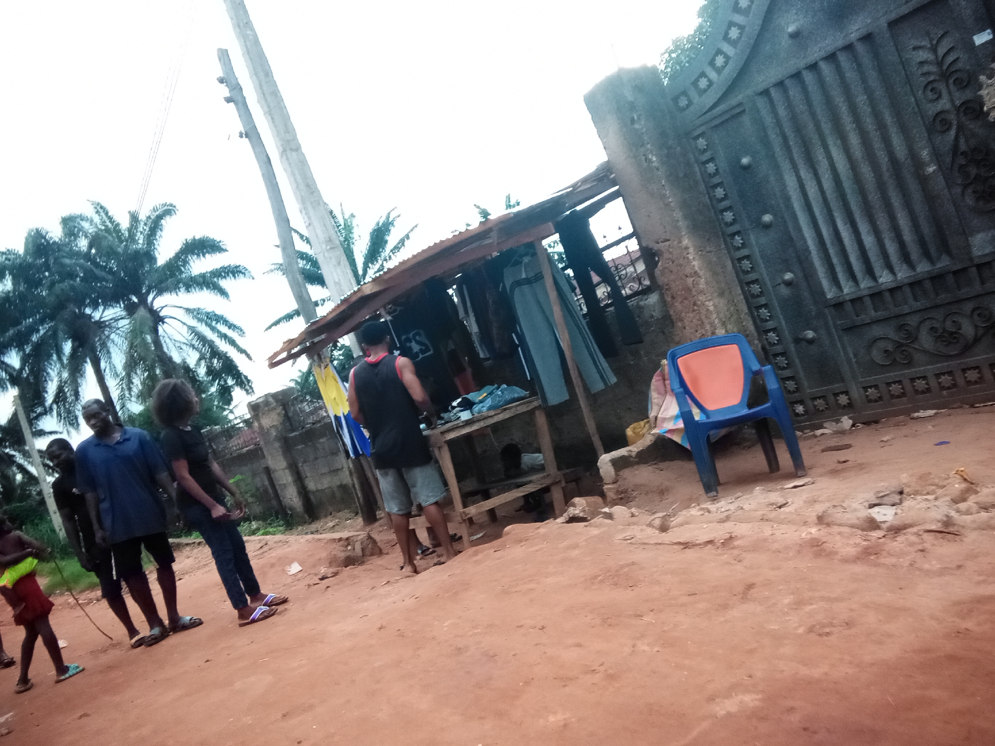 This image captures a street scene in Oko Town, Nigeria. In the foreground, reddish-brown dirt covers the ground, with scattered rocks and debris. Several people are present. To the left, a young child in a red skirt and yellow shirt is walking away from the camera. Behind the child, two adult males stand. One is wearing a dark shirt and dark shorts, and the other, closer to the camera, is wearing a blue polo shirt and dark shorts. To the right of the child, a young woman in a black t-shirt and blue jeans walks away from the camera. In the middle ground, a makeshift stall with a corrugated metal roof is set up. A man in a dark tank top and grey shorts stands facing away from the camera, appearing to be interacting with goods at the stall. Another person is partially visible sitting or lying down inside the stall. Behind the stall, a large, ornate metal gate stands on a concrete pillar. A blue plastic chair with an orange seat is positioned in front of the gate. In the background, tall palm trees are visible against a bright, overcast sky. A wooden utility pole with wires stretches across the upper left portion of the image. The overall impression is a casual, outdoor gathering or market scene in a semi-urban or rural Nigerian setting.