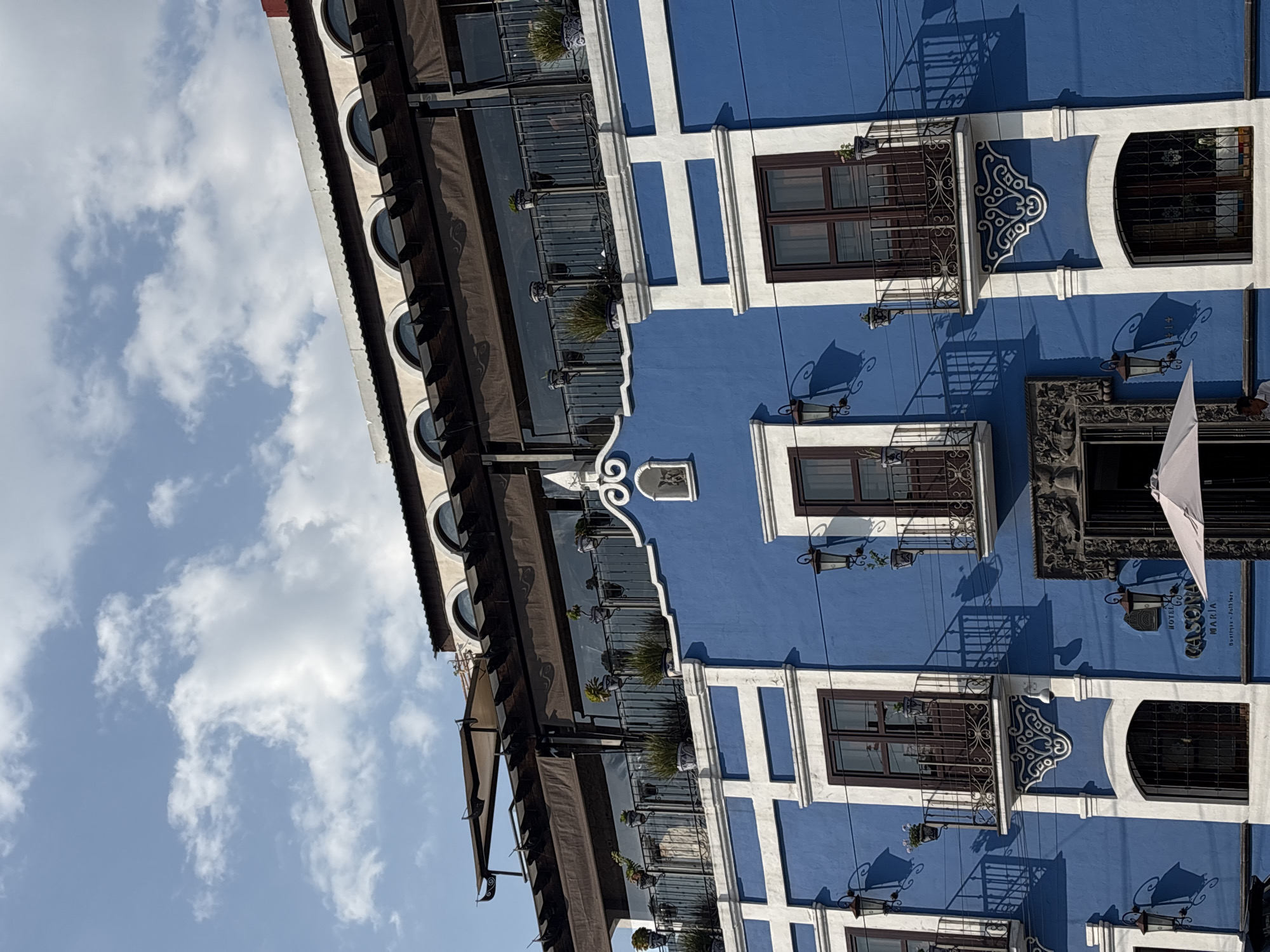The image depicts a section of a bright blue building in Puebla, Mexico. The facade is characterized by white architectural trim that creates a grid-like pattern. The building has a series of windows with dark frames and decorative wrought-iron balconies, some of which feature small potted plants. Additionally, there's a dark awning running along the upper section of the building, with circular window-like openings above it. The sky, filled with scattered clouds, is visible in the upper left of the image. A partially visible sign on the wall reads "HOTEL CASONA MARIA, BOUTIQUE-FOLKLORE". The scene is well-lit, casting shadows on the building's surface.