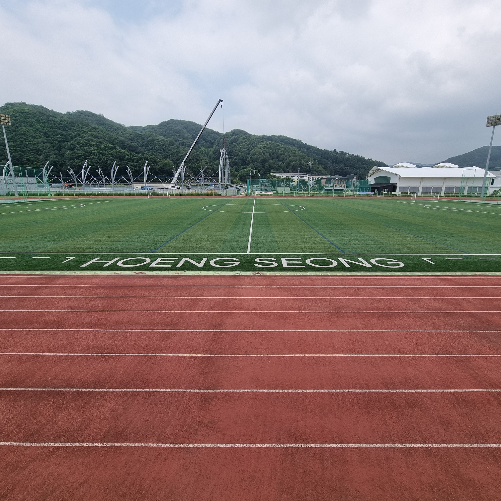 This image captures a wide-angle view of a modern sports complex in Dunnae-myeon, South Korea. In the foreground, a red running track with white lane markings dominates the lower portion of the frame. Beyond the track lies a vibrant green artificial turf field, marked with white lines indicating a soccer pitch, including a central circle and penalty boxes. Along the edge of the turf field, large white letters spell out "HOENG SEONG".
In the background, a scenic mountain range covered in lush green trees rises under a cloudy sky. A large white structure, possibly a gymnasium or arena, with a distinctive curved roof, is visible on the right side of the frame. On the left, a construction crane is actively working on a large, skeletal structure, suggesting ongoing development or renovation within the complex. Other structures, including buildings and stadium lighting, are also present in the distance. The overall scene depicts a well-maintained sports facility situated amidst a natural, mountainous landscape.