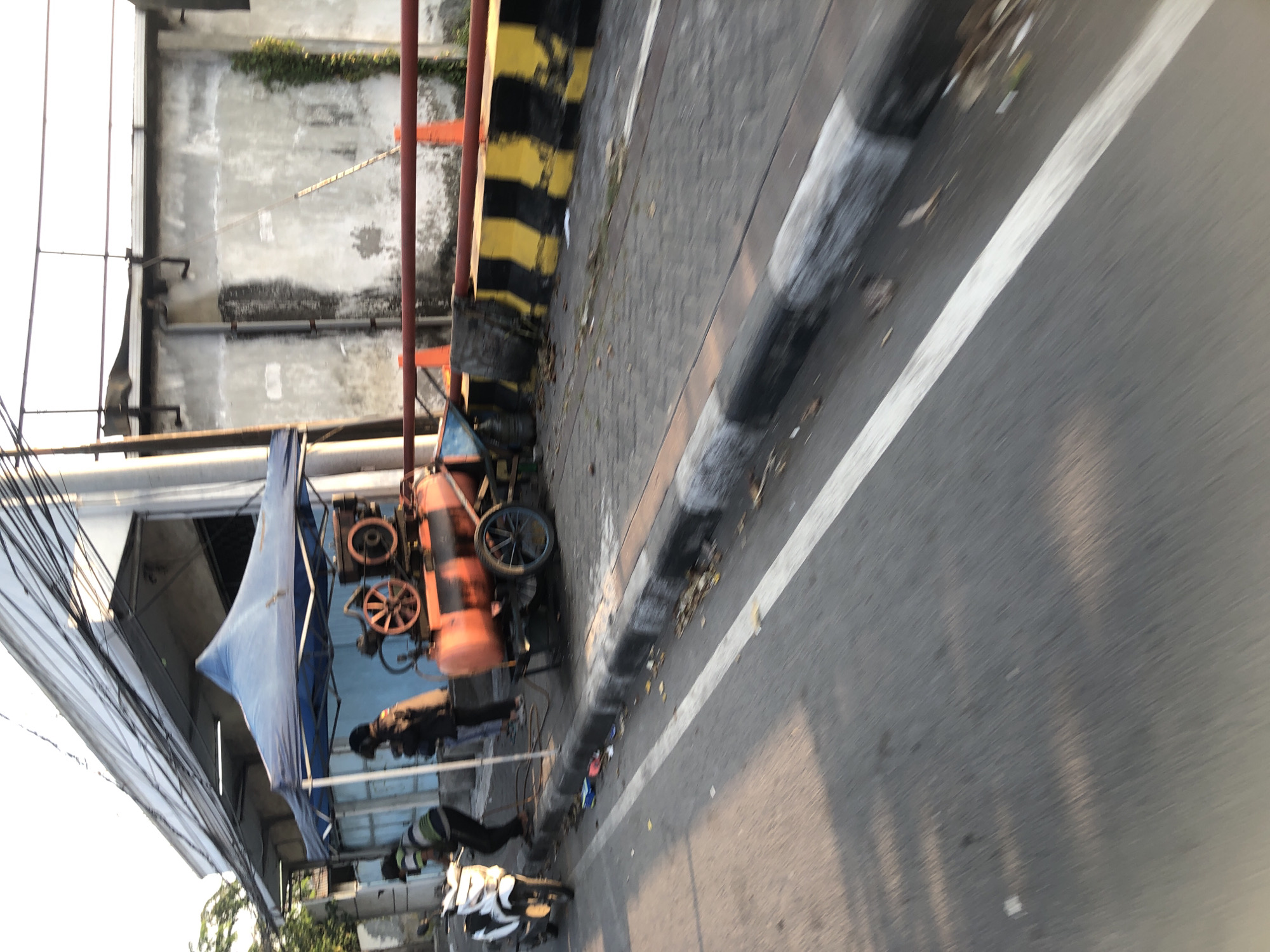 The image depicts an outdoor scene in Gelam, Indonesia, focusing on a street and sidewalk area. A blue tent is set up, partially covering a large, orange industrial compressor with black markings and two wheels. Two individuals, dressed in dark clothing, are near the compressor, with one appearing to be working on it. A white scooter is parked to the left of the tent. The street is paved, with a white line marking a lane. The sidewalk is bordered by a curb with black and white markings, and orange traffic cones are visible further down. The background includes a building with a rough, textured wall and a section of a darker structure. Overhead, power lines are strung across the top of the frame. The lighting suggests it is daytime, with sunlight casting shadows on the road.