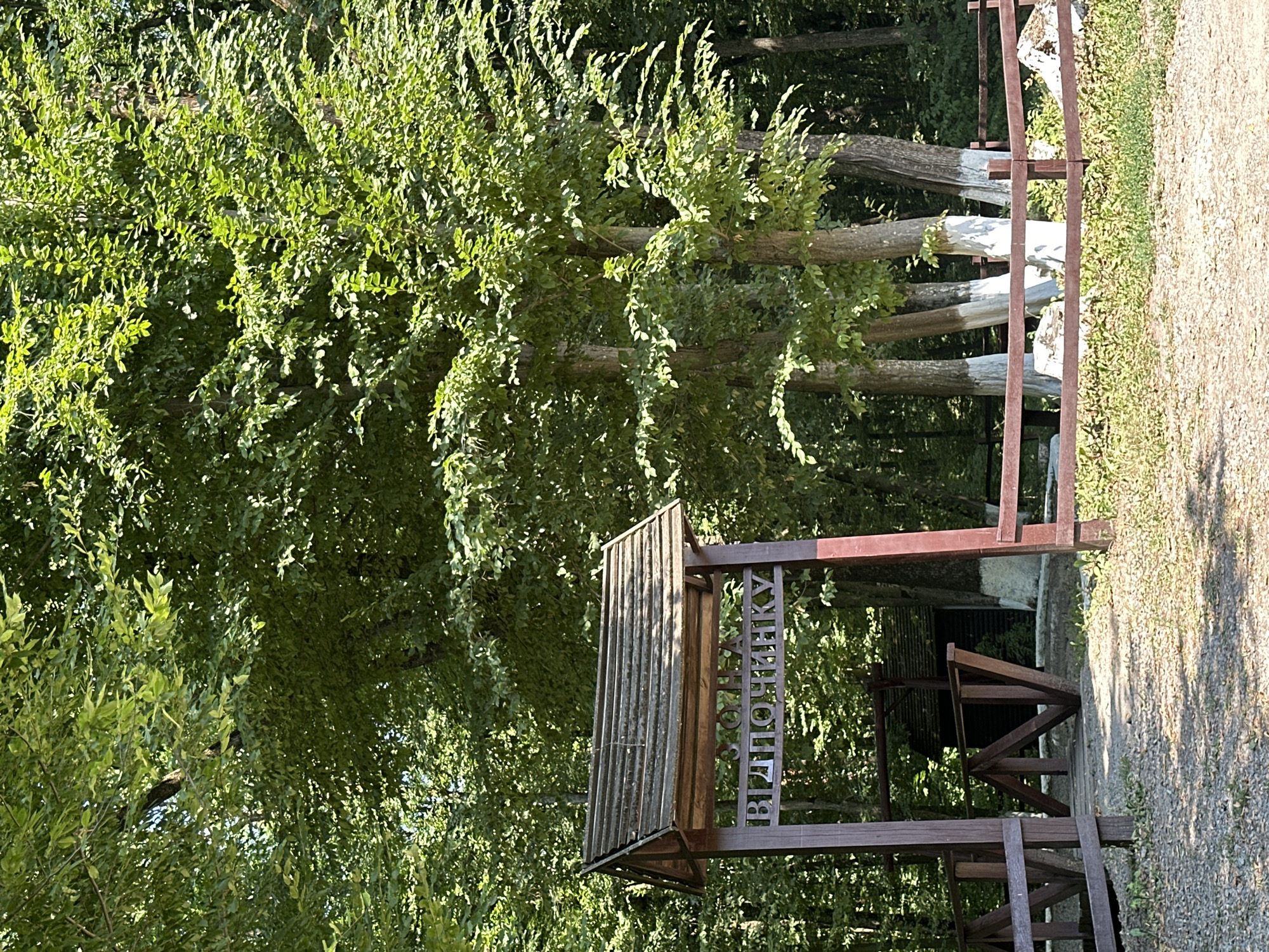 The image shows an outdoor scene with lush green trees and a wooden entranceway. A sign is mounted above the entrance, displaying text in Cyrillic letters. The entrance is bordered by a wooden fence. The ground is covered with gravel and dry leaves. The scene appears to be a park or a natural area. The text on the sign, when translated from Ukrainian, reads "ZONE OF REST". This suggests the location is a designated area for relaxation within a park or recreational space. The presence of trees and the overall natural setting indicate it is an outdoor environment. The overall impression is one of a serene and natural setting, possibly a park or a nature reserve.