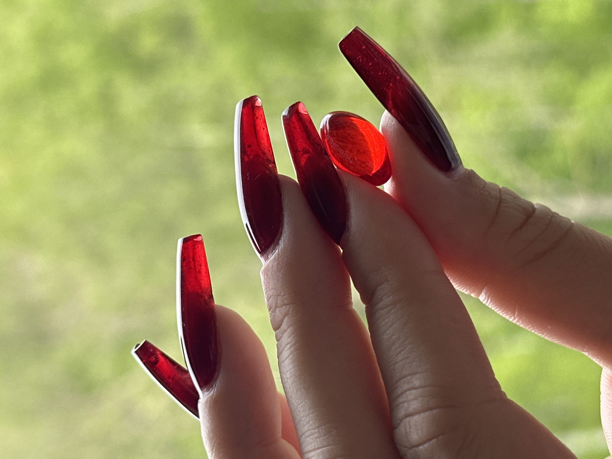 A close-up photograph shows a hand with long, red, acrylic nails holding a red, translucent capsule pill. The nails are rectangular in shape and appear to be partially transparent. The background is a blurred, green scene, possibly indicating an outdoor location with foliage. The hand is light-skinned and the focus is on the nails and the pill. The location is unknown.