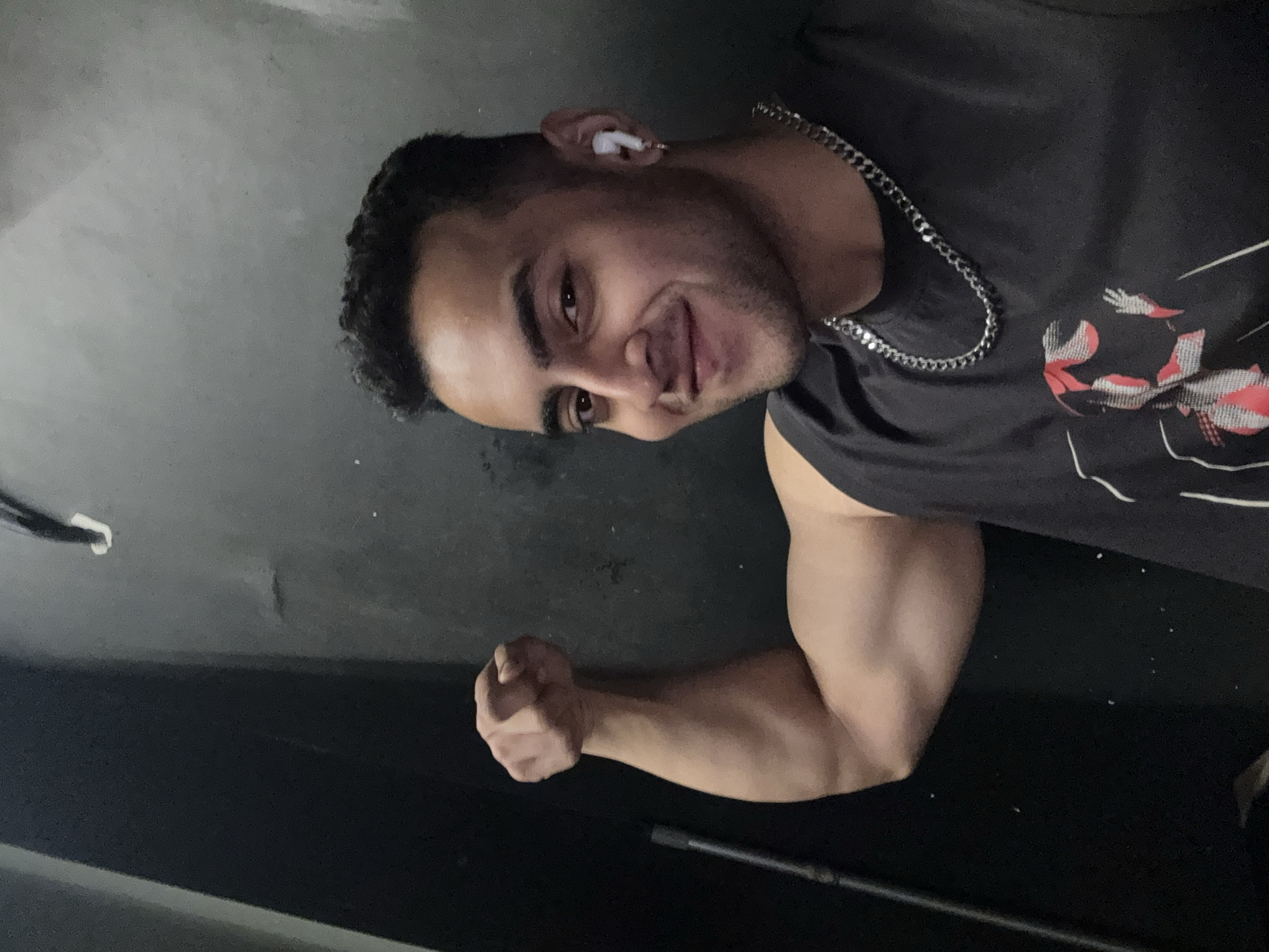 A man is posing in a room. He has short, dark hair, a chain necklace, and an earbud in his left ear. He is wearing a gray sleeveless shirt with a design. He is flexing his right bicep and smiling at the camera. The room has a dark, possibly black, wall and floor. There is a cord visible in the upper left corner and a black rod on the floor. No information indicates that this picture was taken in Kolkata, India.