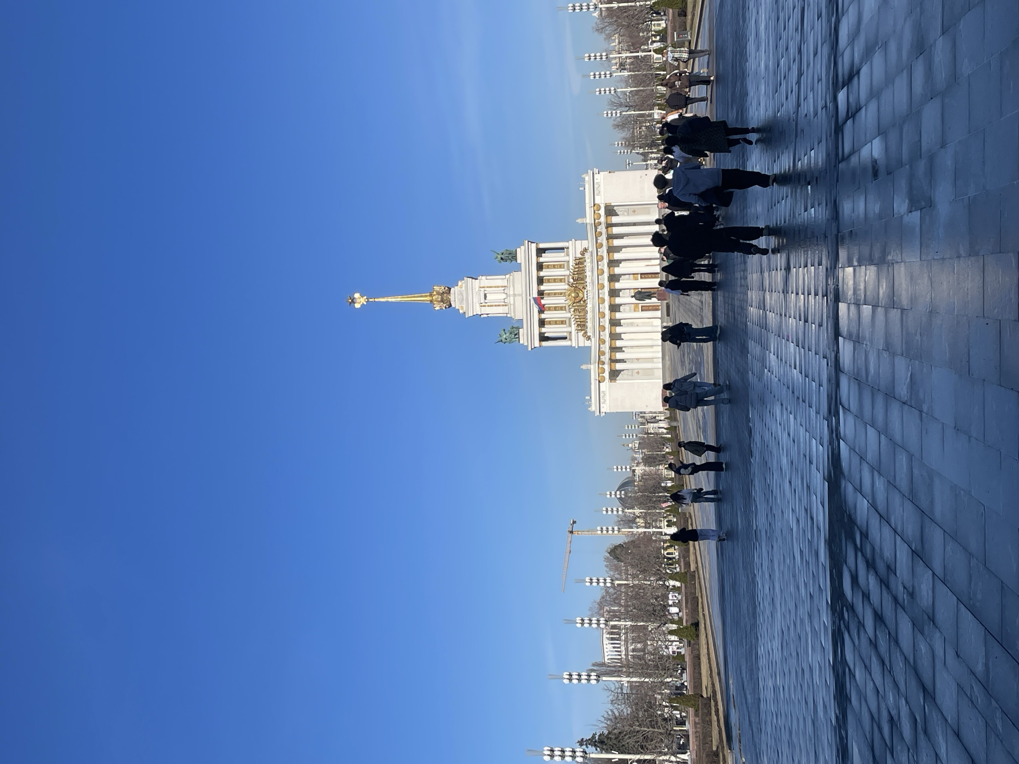 A low-angle, eye-level shot shows a white, multi-tiered building with gold accents and a spire topped with a five-pointed star. The building is located in the city of Khimki, Russia. Several people are walking on a paved area in front of the building. The sky is clear and blue. Streetlights line the walkway. A construction crane can be seen in the background.