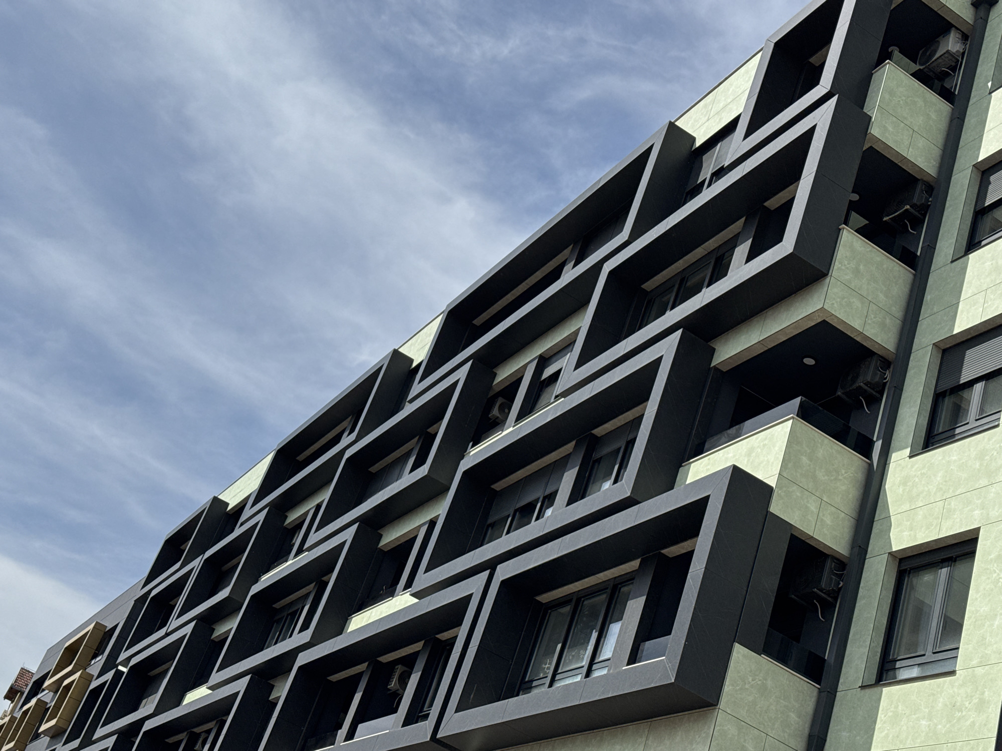 A low-angle medium shot captures a modern apartment building in Novi Sad, Serbia. The building features a unique exterior design, with dark, box-like structures framing the windows and balconies, set against a light green facade. The sky is visible in the background, exhibiting a cloudy pattern. Air conditioning units are visible on some of the balconies. The overall architectural style is contemporary.