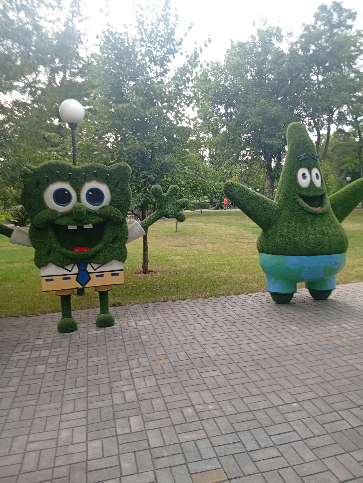 In Pokrov, Ukraine, two topiary figures resembling cartoon characters Spongebob Squarepants and Patrick Star stand on a paved walkway in a park. Spongebob, on the left, is depicted as a green, square-shaped figure with large blue eyes, buck teeth, and a white shirt with a blue tie and yellow suspenders. Patrick, on the right, is a green, star-shaped figure with wide eyes and a smiling mouth, wearing blue shorts. Both figures are made of green shrubbery and are positioned outdoors amidst lush green trees and grass. The sky is overcast, suggesting a cloudy day. The paving stones are arranged in a herringbone pattern.