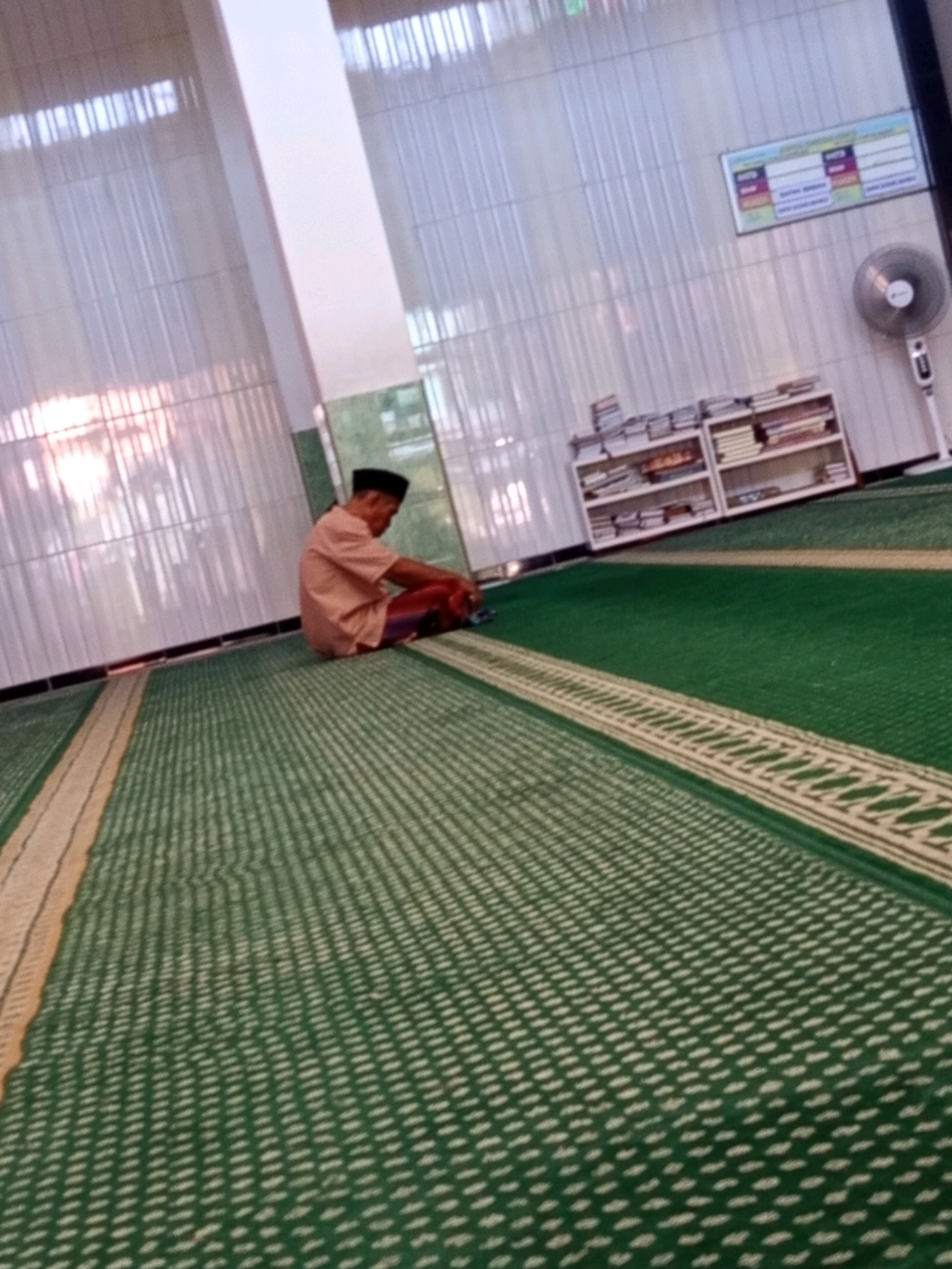 A man wearing a light orange shirt and a dark cap sits on a green carpeted floor in what appears to be a mosque. He is facing away from the camera and is positioned in the middle ground, to the left of the frame. The carpet has a pattern of small, light-colored dots and a decorative border. In the background, there are white shelves filled with books, a ceiling fan, and a framed schedule on the wall. The overall environment suggests a place of worship or study. The image does not provide specific details to pinpoint the exact location within Karawang, Indonesia.