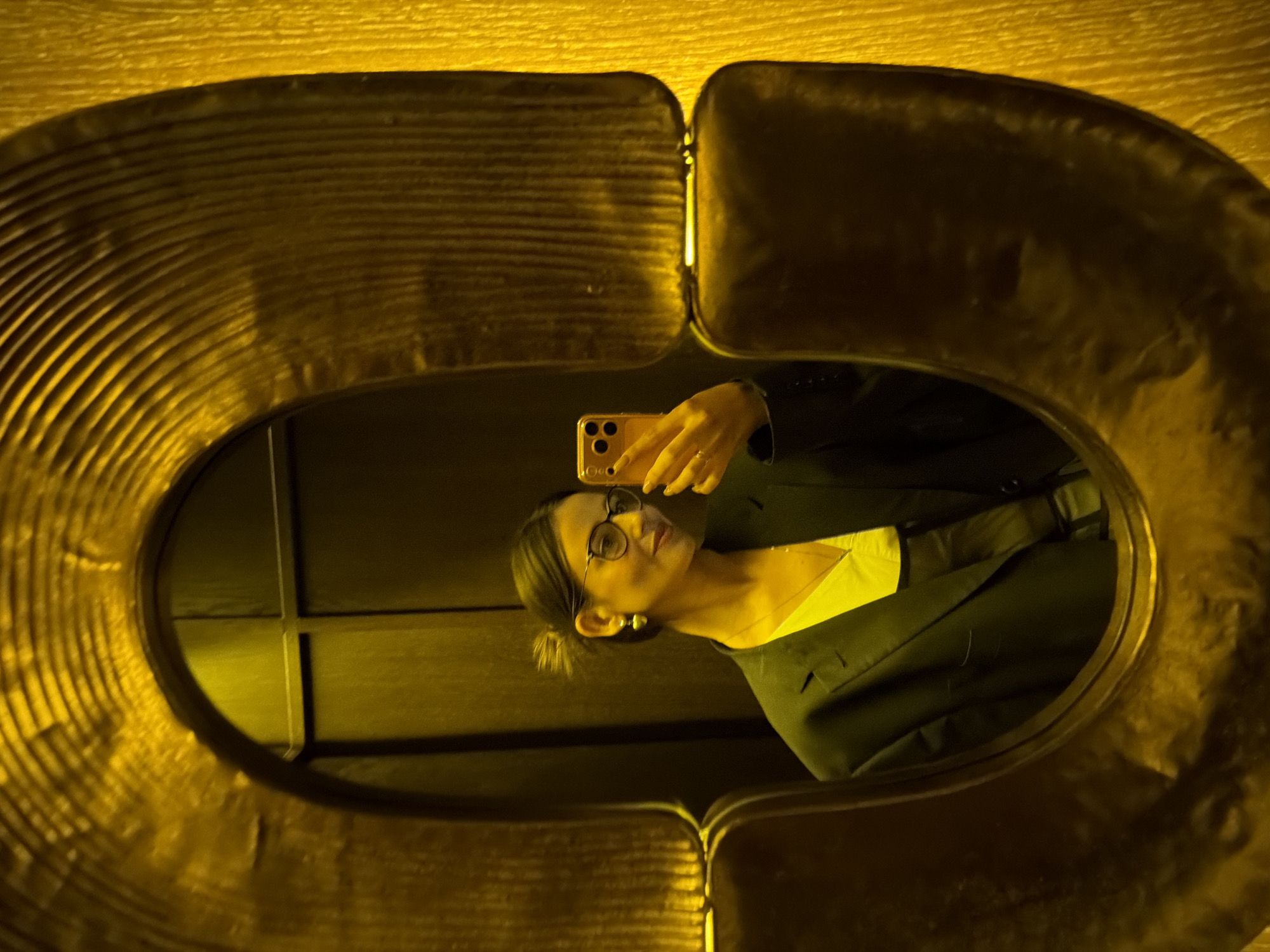 This media file is a photograph taken in Krasnoyarsk, Russia. It shows a reflection of a person in a decorative, oval-shaped mirror. The person, likely a woman, is wearing glasses, a dark blazer over a bright yellow top, and large earrings. She is holding a smartphone in her right hand, appearing to take a selfie. The background reflected in the mirror seems to be a dimly lit room with wooden paneling. The mirror itself has a textured, golden frame that creates a stylized border around the reflection. The lighting in the image is warm and yellow-toned, suggesting artificial light, possibly in the evening or a dimly lit indoor space. The overall mood is casual and personal, as is typical of a selfie. There is no visible text in the image.