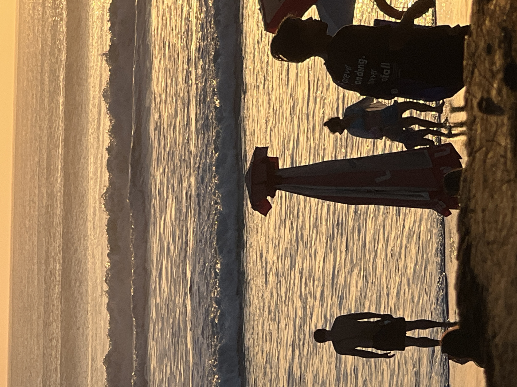 In Kuta, Indonesia, at sunset, several individuals are silhouetted against the shimmering ocean. A man stands facing the water, his back to the camera. To his right, a beach umbrella is prominent. Further to the right, a group of people, including a child, are partially obscured by another umbrella. In the foreground, a dark silhouette of a person with text on their shirt faces away from the camera, looking out at the water. The ocean features gentle waves reflecting the warm golden light of the setting sun, and the sky above is a soft, hazy orange.