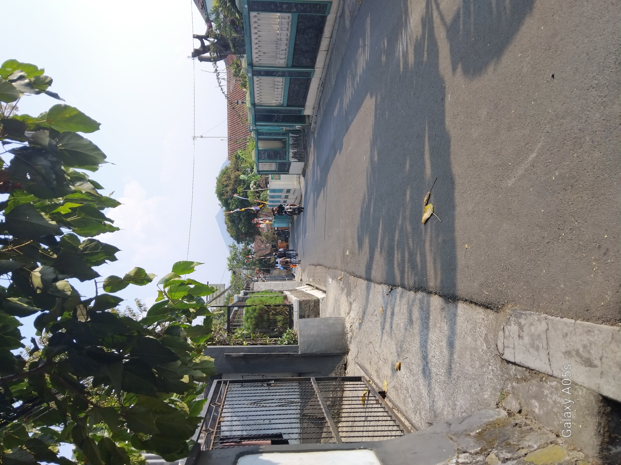 The image captures a sunlit residential street in Sumedang, Indonesia. A paved asphalt road occupies the center, with a concrete sidewalk running alongside it on the left. Lush green foliage from overhead trees casts dappled shadows across the road and sidewalk. On the left, a black metal gate provides access to a property with a dark gray wall. The right side of the street is lined with a decorative fence featuring a light-colored upper section with arched patterns and a darker lower section. Several houses with tiled roofs are visible behind the fence. In the distance, a mountain peak is discernible against a bright, clear sky. Two individuals are riding bicycles on the road, and a person on a motorcycle is also present. Fallen leaves are scattered on the road and sidewalk. The scene conveys a quiet, daytime ambiance in a suburban or semi-rural area of Sumedang.