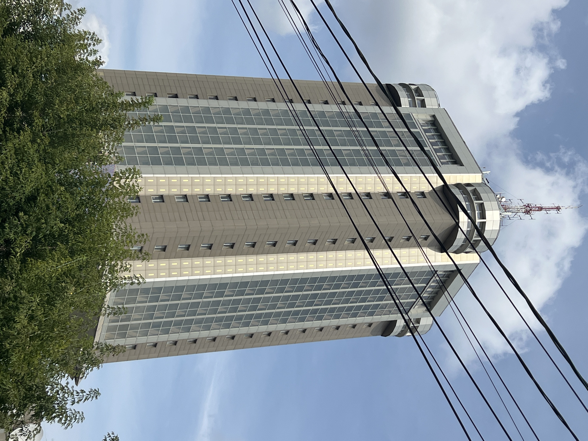A tall, modern skyscraper stands against a partly cloudy blue sky in Tver, Russia. The building's facade is a combination of light brown panels and large, grid-like windows, with some sections featuring vertical strips of what appear to be illuminated elements. The top of the skyscraper is crowned with a complex antenna array. In the foreground, lush green trees partially obscure the lower portion of the building, and several dark electrical wires crisscross the frame in the mid-ground, creating a layered perspective. The scene is static and depicts an urban architectural element in its environmental context.