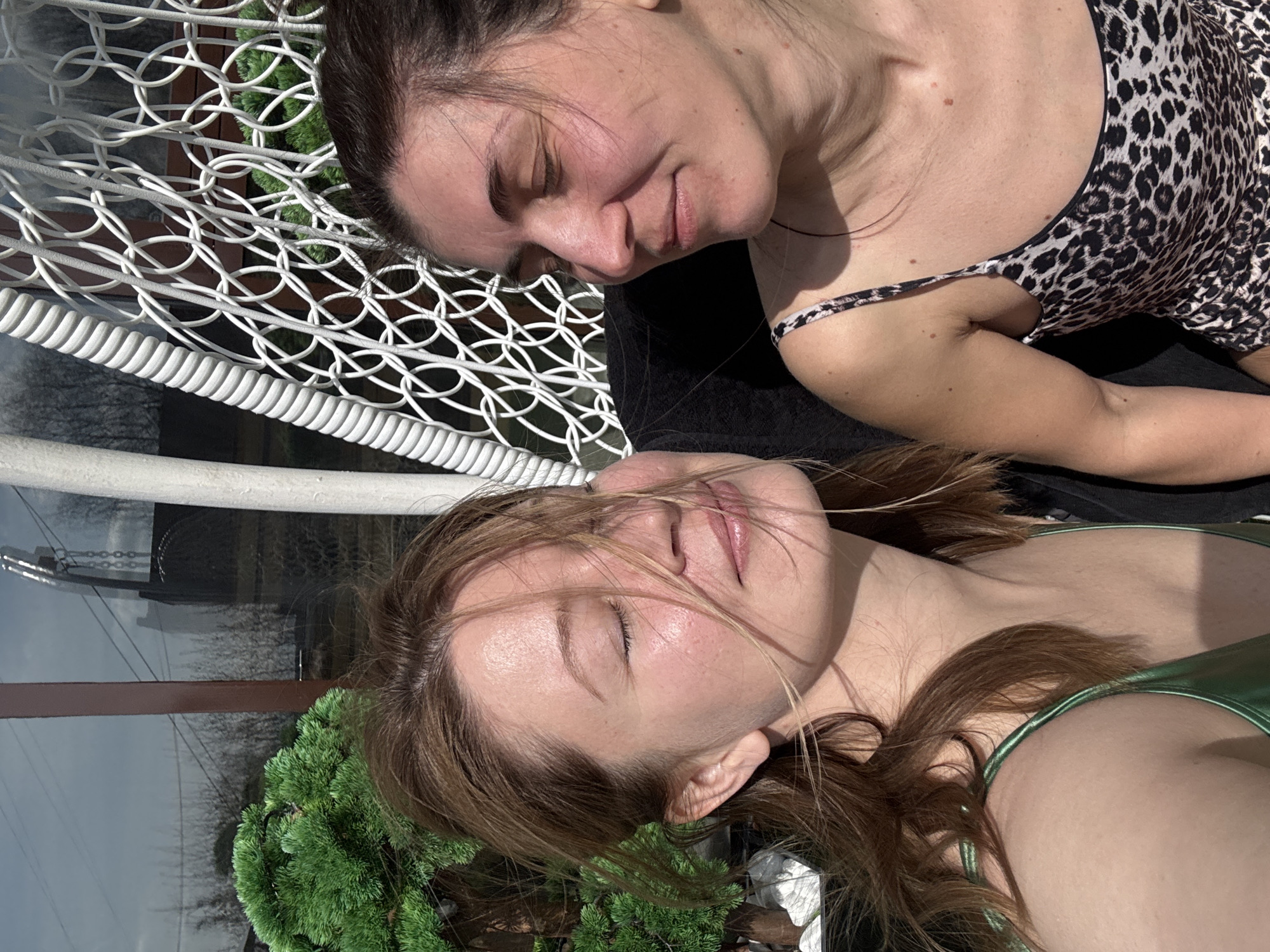 Two women are shown in a close-up, overhead shot, seemingly resting or sunbathing outdoors in Votkinsk, Russia. Both are lying down with their eyes closed, indicating a state of relaxation or sleep.
The woman in the upper part of the frame has dark brown hair, pulled back, and is wearing a leopard print top with thin straps. Her face is turned slightly towards the viewer, showing a soft, contented expression with flushed cheeks. Her left arm is partially visible.
The woman in the lower part of the frame has lighter brown hair, with some strands gently falling across her face. She is wearing a shiny green top, also with thin straps. Her face is turned towards the viewer, revealing a peaceful expression with slightly parted lips. Her right shoulder and arm are visible.
Both women appear to be lying on a dark, possibly black, surface. To the left of the image, a prominent white lattice-like structure, possibly part of a garden swing, hammock, or decorative screen, provides a patterned backdrop. Below the lattice, a thick, white, rope-like structure is visible. Behind the lower woman's head and in the bottom left, dense green foliage, resembling a small bush or tree, adds a natural element to the scene. In the background, behind the foliage, there's a faint reflection that suggests a building or window under a cloudy sky.
The lighting is bright and sunny, indicating daytime, with strong highlights on the women's skin and hair. The overall mood of the image is serene and relaxed, capturing a moment of calm rest. No visible text is present in the image.
