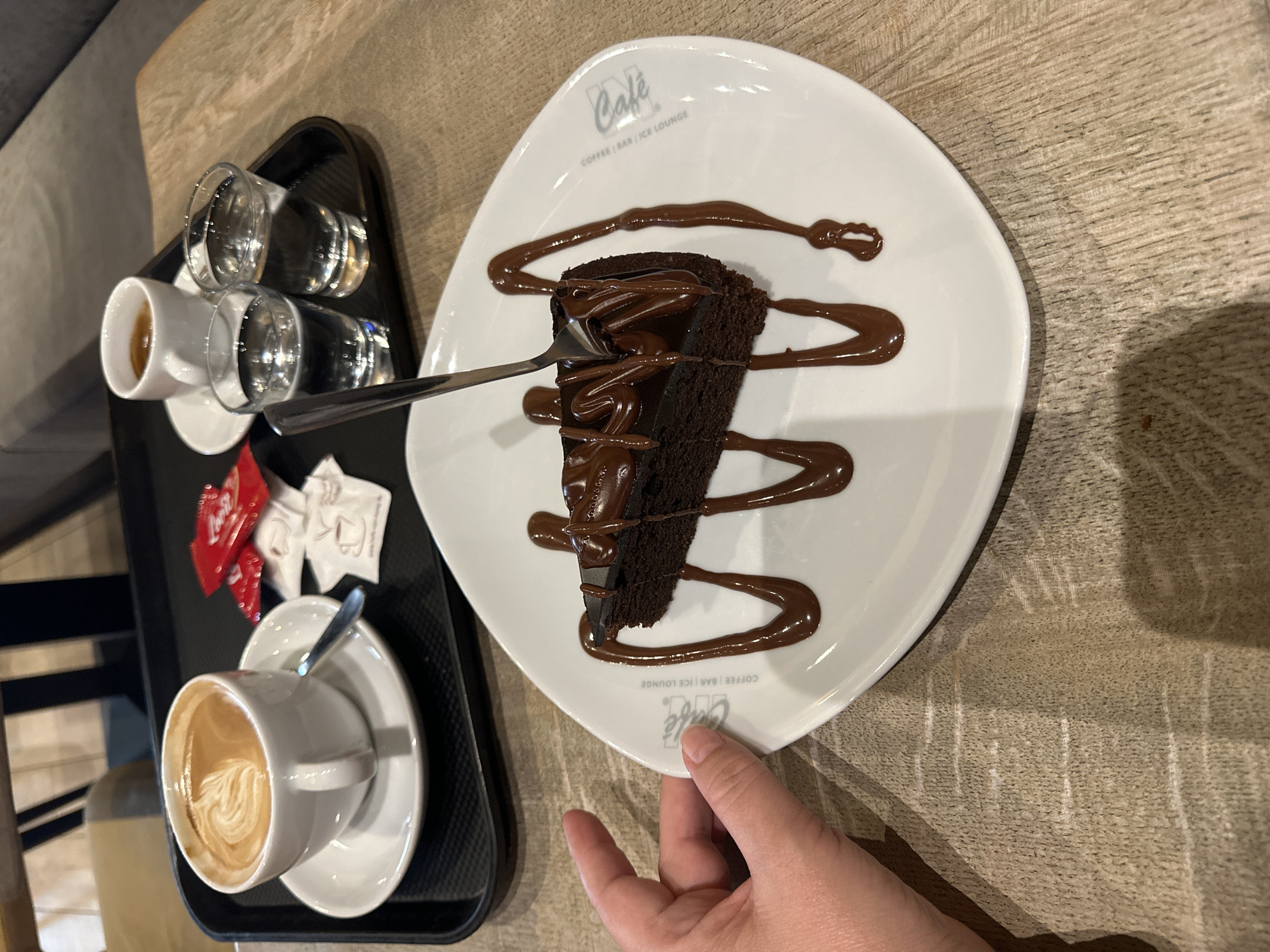 A slice of chocolate cake drizzled with chocolate sauce sits on a white plate with the cafe logo. The plate is on a wooden table, next to a tray holding two small glasses of water, a small cup of espresso, and a latte macchiato. A hand is visible on the left side of the image, reaching towards the plate. The cafe is identified as "Cafe - Coffee | Bar | Ice Lounge" in Krefeld, Germany.