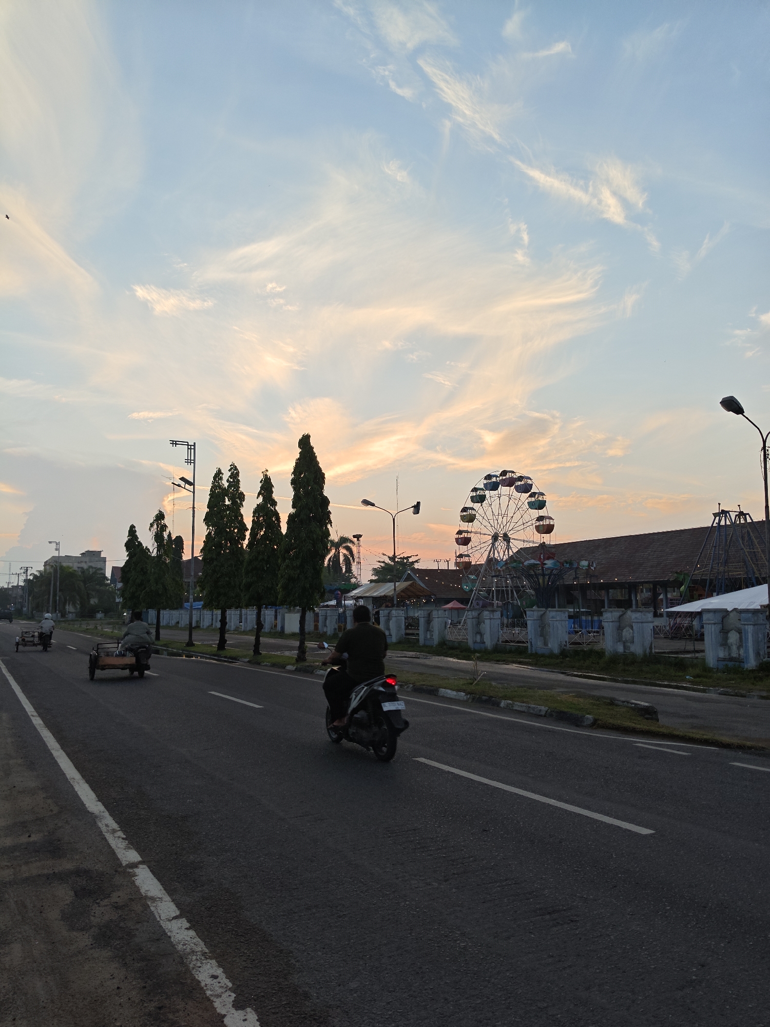 The image captures a street scene in Lhokseumawe, Indonesia, likely during the late afternoon or early evening, given the soft, warm light and wispy clouds in the sky. The sky is a blend of pale blue and orange hues, suggesting the sun is low on the horizon. A Ferris wheel, brightly colored with multiple gondolas, stands prominently in the background, indicating the presence of an amusement park or fairground. Adjacent to the Ferris wheel are several low-rise buildings with dark roofs, possibly stalls or structures associated with the park.
In the foreground, a paved road stretches into the distance, marked with white lines. Several vehicles are visible on the road. Two motorbikes, each carrying a rider, are traveling away from the viewer. One motorbike has a sidecar attached. Further down the road, a cart-like vehicle, also with a rider, is moving in the same direction.
Lining the left side of the road are several tall, slender, dark green trees, forming a visual barrier. Beyond the trees, taller buildings are partially visible, suggesting a developed urban area. Streetlights are positioned along the road, and one lamp post is particularly prominent, equipped with solar panels, hinting at sustainable energy initiatives. The overall atmosphere is tranquil and somewhat hazy, typical of a tropical environment. The presence of the Ferris wheel and amusement park elements suggests this area is a recreational hub within Lhokseumawe.