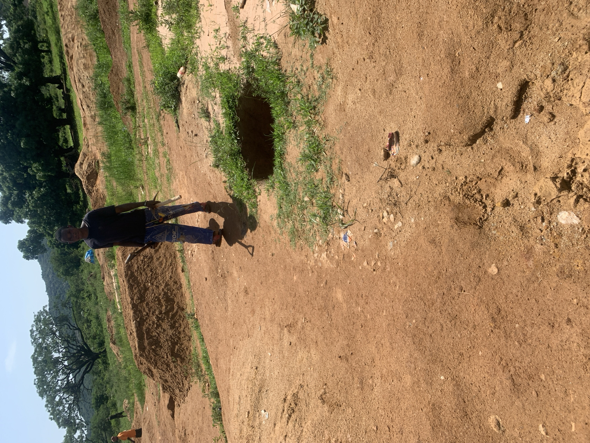 In Buga da Gindi, Nigeria, a young individual stands in a dusty, open area. They are wearing a dark t-shirt and blue pants with a white pattern. The individual is holding a shovel with a wooden handle and a white end. The environment is characterized by piles of brown dirt, sparse green grass, and a clear sky. In the background, there are large trees and hills. To the left of the individual, another person in an orange shirt is visible, also near a pile of dirt. A hole is present in the ground to the right of the standing individual. There is scattered debris on the ground, including a white plastic bottle and what appears to be a discarded shoe. The ground is predominantly reddish-brown earth.