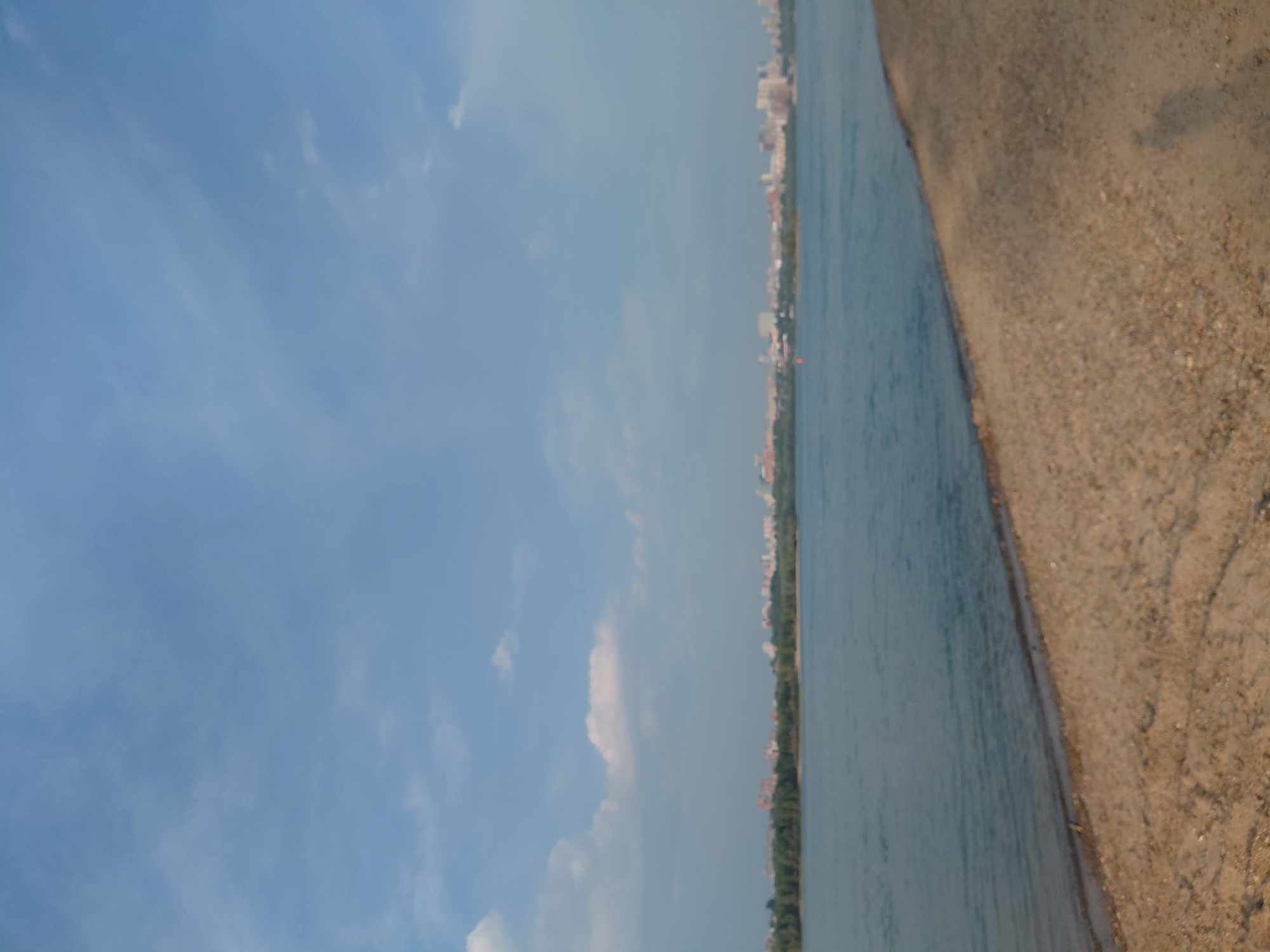 The image depicts a wide river with a sandy embankment in the foreground. In the distance, across the river, lies the city of Novosibirsk, Russia. The skyline is characterized by buildings of various heights, interspersed with trees along the riverbank. The sky above is partly cloudy, with a mix of blue sky and white clouds. The water appears calm and reflects the colors of the sky and the distant city. A shadow of a person is visible on the sand in the lower right portion of the image, suggesting the photographer's presence. The overall scene conveys a peaceful, urban riverside landscape.