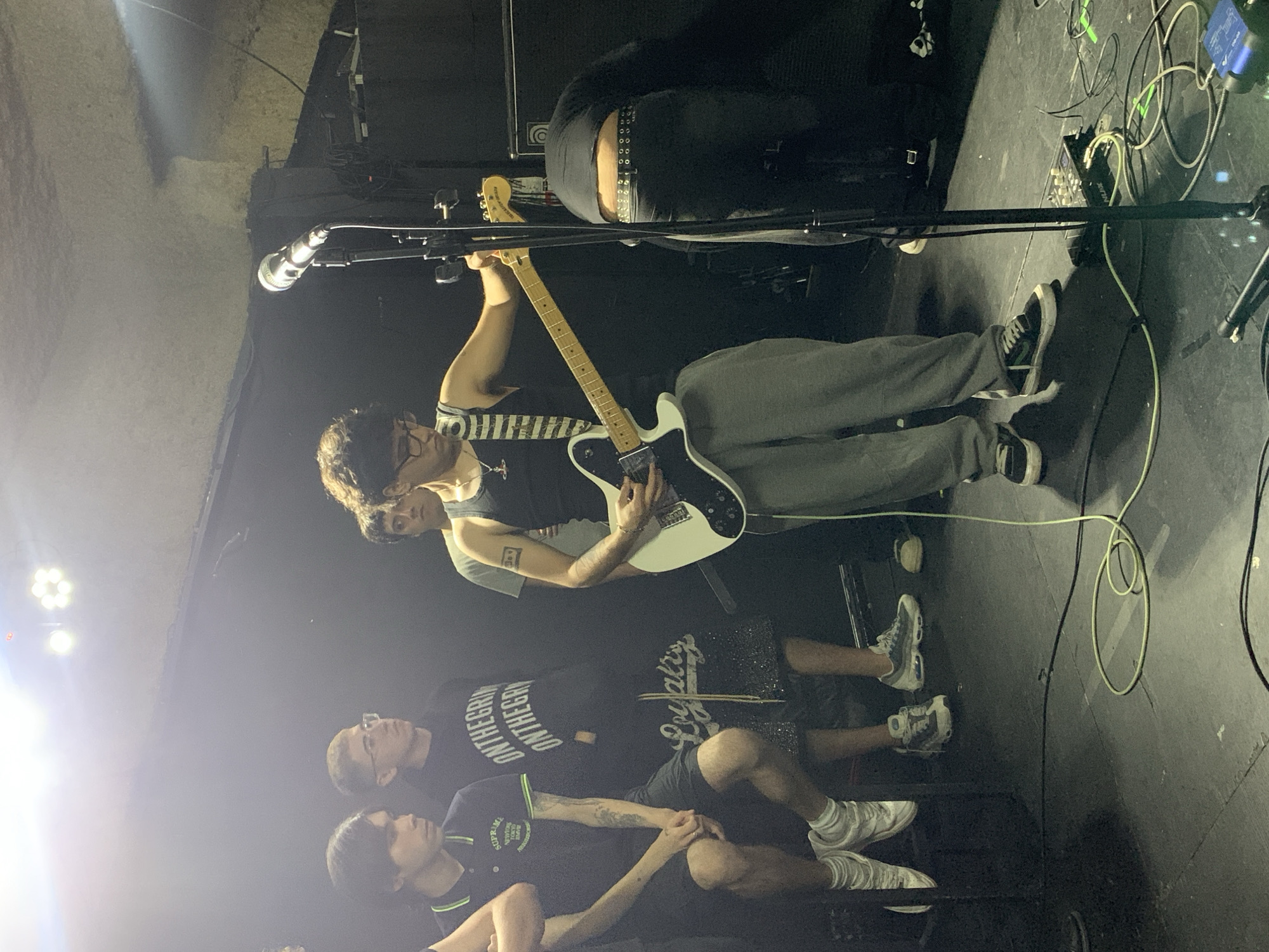 A band performs on a dimly lit stage. A guitar player with glasses is playing a white electric guitar while wearing a dark tank top and gray sweatpants. A microphone stands in front of him. Below and slightly to the left of the guitar player, three people sit on the edge of the stage, seemingly watching the performance. One of them wears shorts with the word "Loyalty" on them. Stage lighting illuminates the left side of the image. Cables and equipment are scattered on the floor. It's likely an underground music venue in Buenos Aires, Argentina.