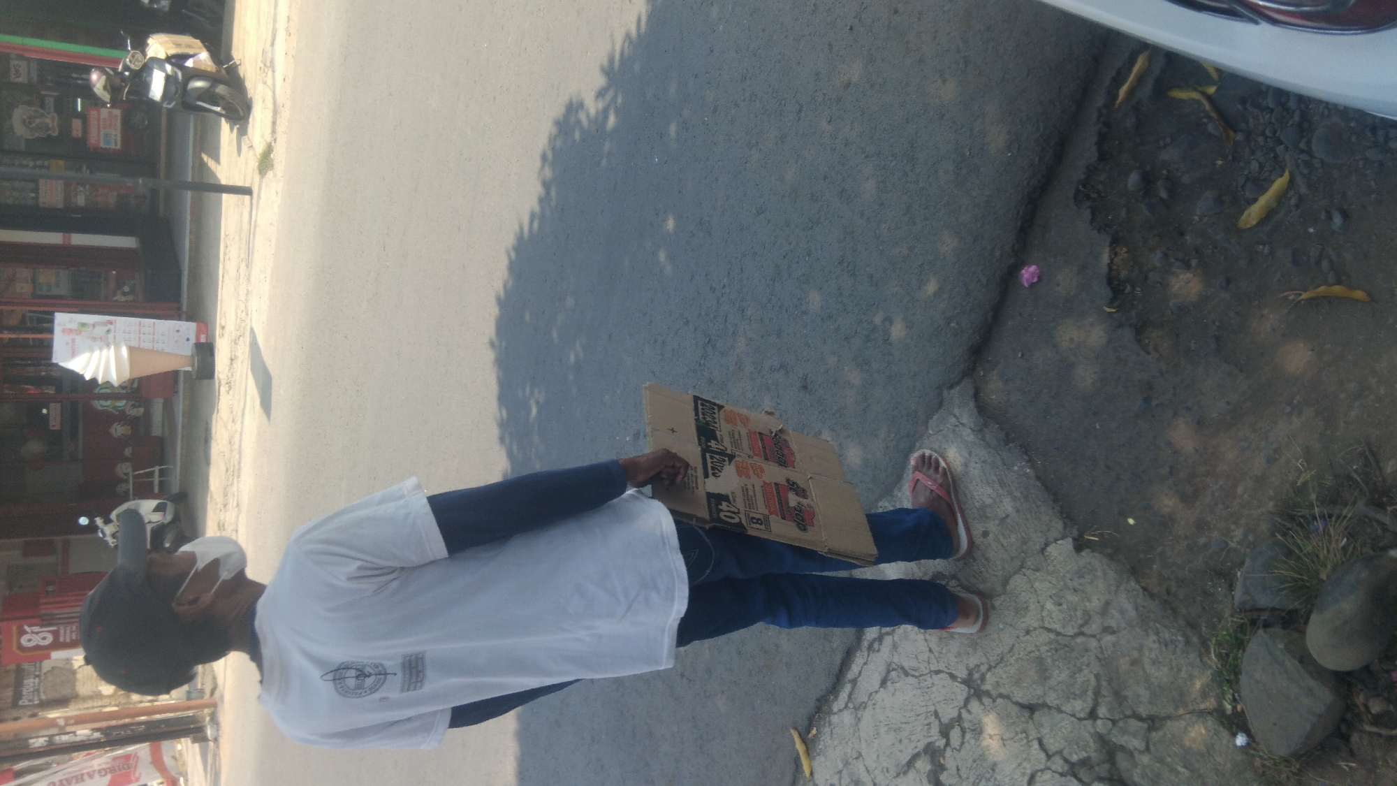 A person wearing a white t-shirt, blue pants, a black cap, and a white face mask stands on a cracked concrete sidewalk. They are holding a piece of corrugated cardboard in their right hand. The cardboard has red and black text and graphics printed on it. The person is facing away from the camera, looking towards a paved road. In the background, across the road, is a storefront with a large ice cream cone statue outside. A white motorcycle with a yellow box on the back is parked in front of the storefront. Sunlight casts shadows on the road. The environment appears to be an urban or commercial area.