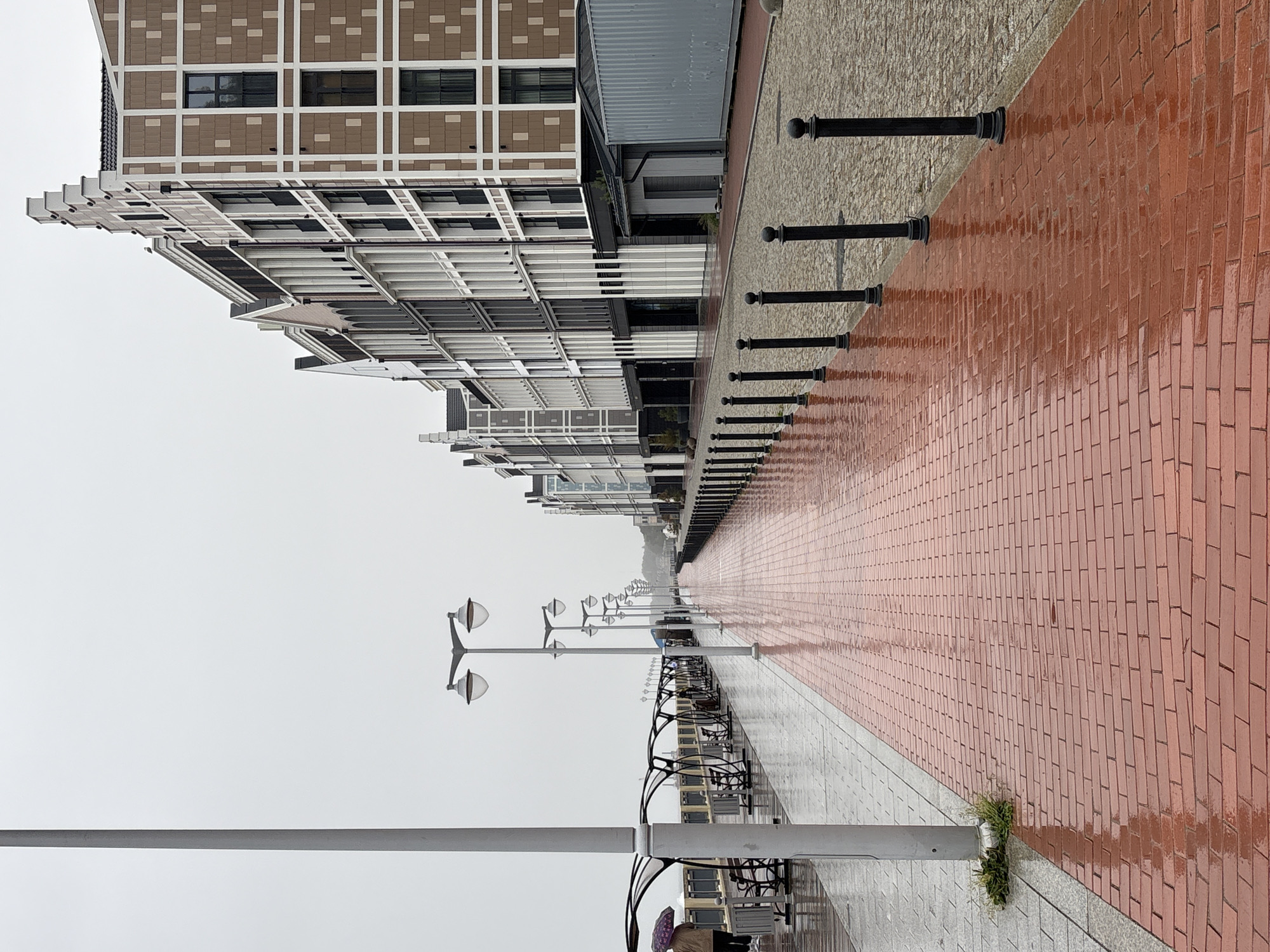 The image depicts a waterfront promenade in Svetlogorsk, Russia, on a cloudy day. The promenade is paved with red bricks, which are wet and reflective from rain. On the left side of the promenade, there are covered benches and a tall lamppost. A person holding a purple umbrella is visible walking away from the camera on the left. To the right of the promenade, a row of black bollards separates it from a cobbled street. Beyond the street, a series of modern buildings with distinctive architectural features, resembling Dutch gables, line the promenade. The sky is overcast and white.