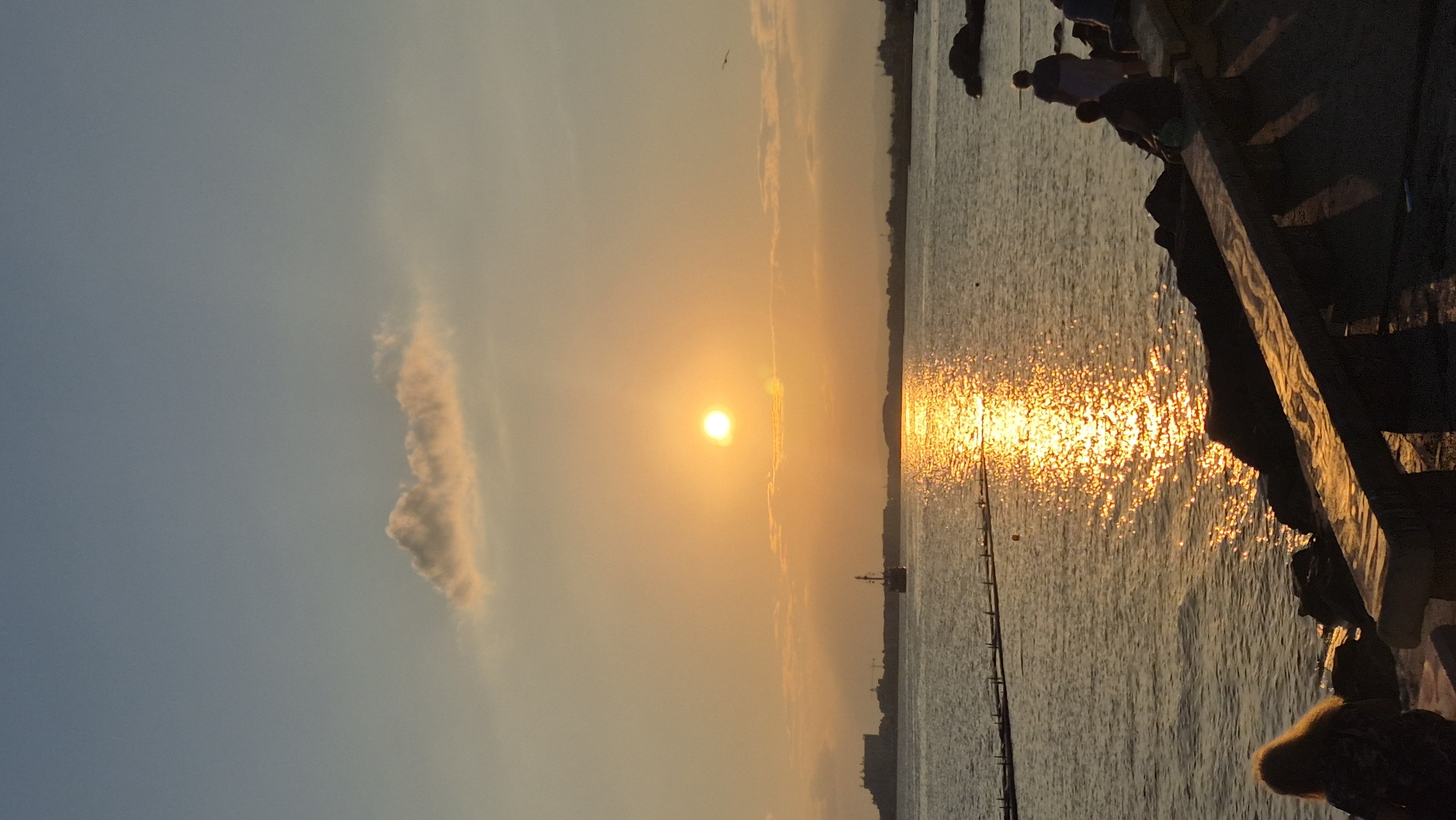 The image depicts a sunset over the Black Sea in Pomorie, Bulgaria. The sun, a bright orb, is positioned near the center of the frame, casting a golden, shimmering reflection on the water. A small boat is visible in the distance, sailing across the reflective surface. The shoreline in the background shows a silhouette of buildings and hills. In the foreground, several people are seated or standing on a rocky embankment, observing the sunset. One person with long blonde hair is partially visible on the left. The sky transitions from a pale blue at the top to warm orange and yellow hues near the horizon.