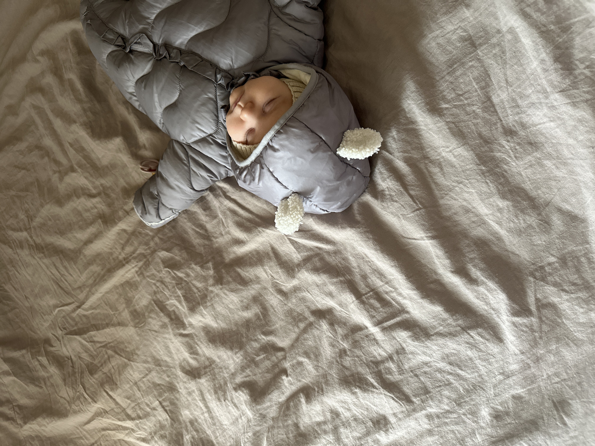 A baby sleeps in a gray, quilted snowsuit with white pom-poms on a tan-colored bedsheet. The location is not discernible from the image.