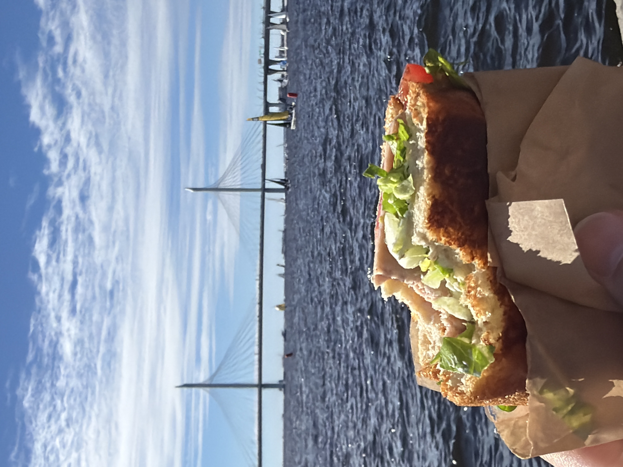 A close-up shot of a partially eaten sandwich held in a person's hand, with the ZSD Bridge in St. Petersburg, Russia, and the Gulf of Finland in the background. The sandwich is wrapped in brown paper and filled with lettuce, tomato, and meat. The bridge, a cable-stayed bridge, spans a wide body of water. Several sailboats are visible on the water, indicating a recreational activity. The sky is blue with scattered white clouds. The image appears to be taken on a sunny day.