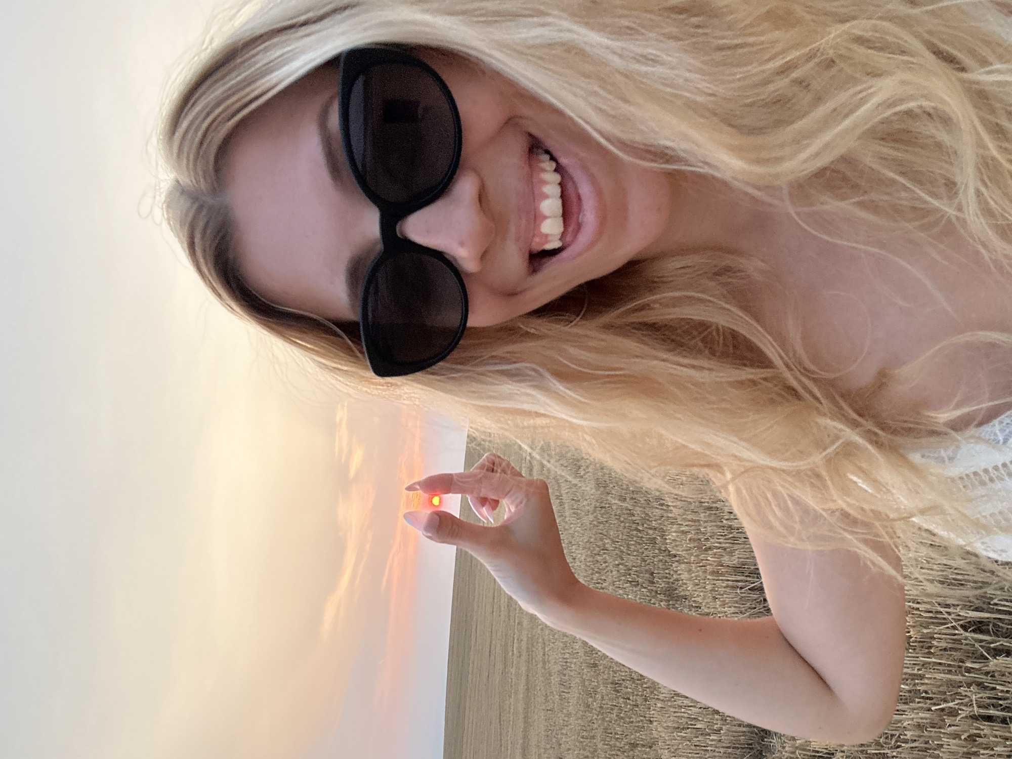 The image shows a young woman with blonde hair and sunglasses taking a selfie in a harvested field at sunset. She is holding up her hand to frame the setting sun between her thumb and forefinger. The sky is filled with soft orange and pink hues, and the field is covered with dry stubble. The woman is smiling and sticking out her tongue.