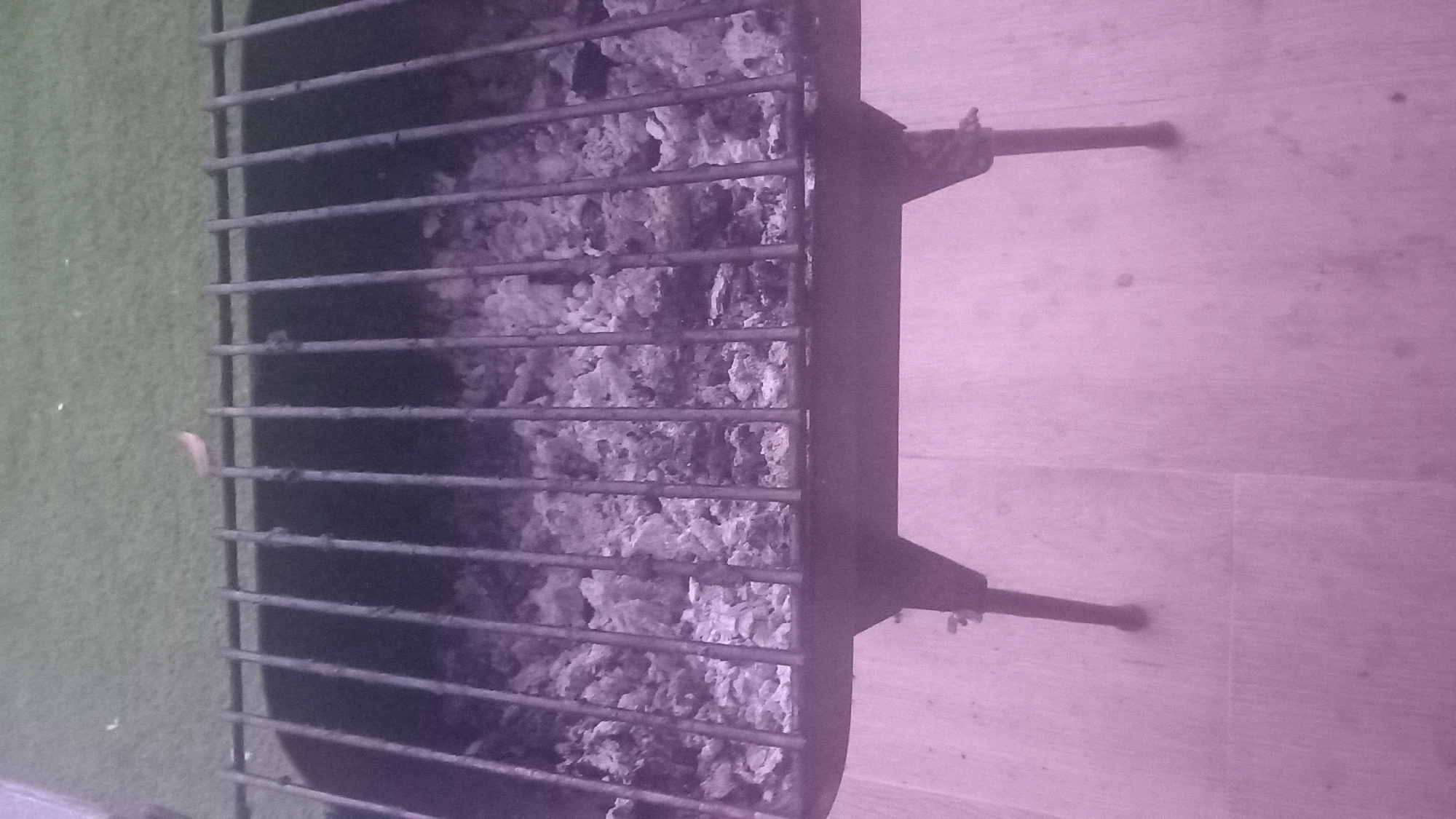 The image shows a close-up, rotated view of a barbecue grill. The grill is made of dark metal bars, and underneath the bars, there are remnants of ash and charcoal, indicating it has been used recently. The grill has two legs visible on the right side, propping it up. To the left of the grill, a patch of green, possibly artificial turf or a mat, is visible. The background consists of a light-colored wooden surface, likely a floor or tabletop, with faint grain patterns. The lighting in the image has a strong purple hue, suggesting artificial lighting or a color filter. No people are present in the image. The scene is likely outdoors or in a covered outdoor area given the presence of a grill and turf. There are no visible text elements in the image.